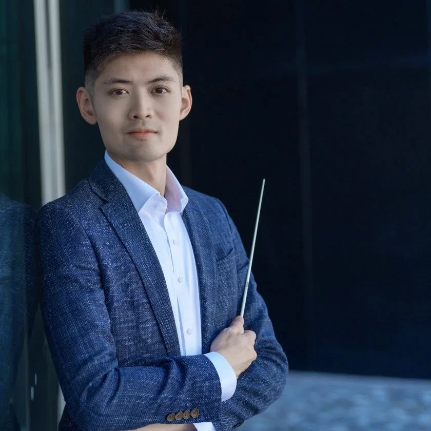 A young man in a blue blazer and white shirt standing against a dark background, looking at the camera, holding a thin pointing stick.
