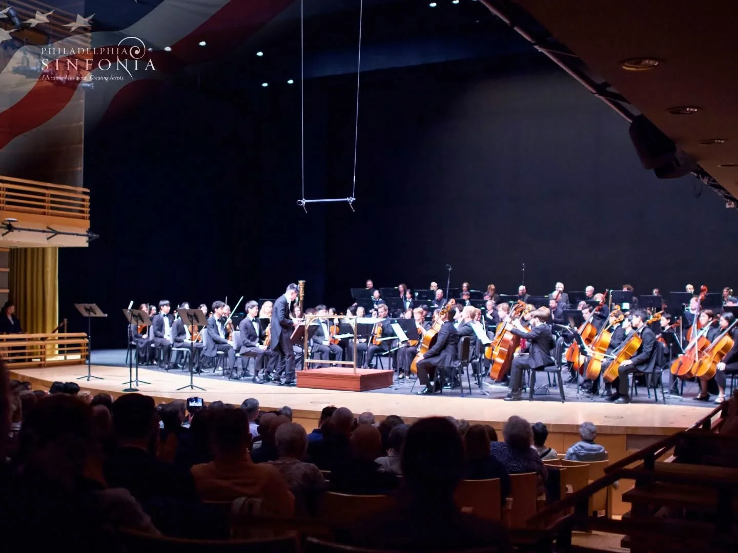 Orchestra performing on stage in a concert hall with audience seated in front. Musicians are playing string instruments, and a conductor is leading them.