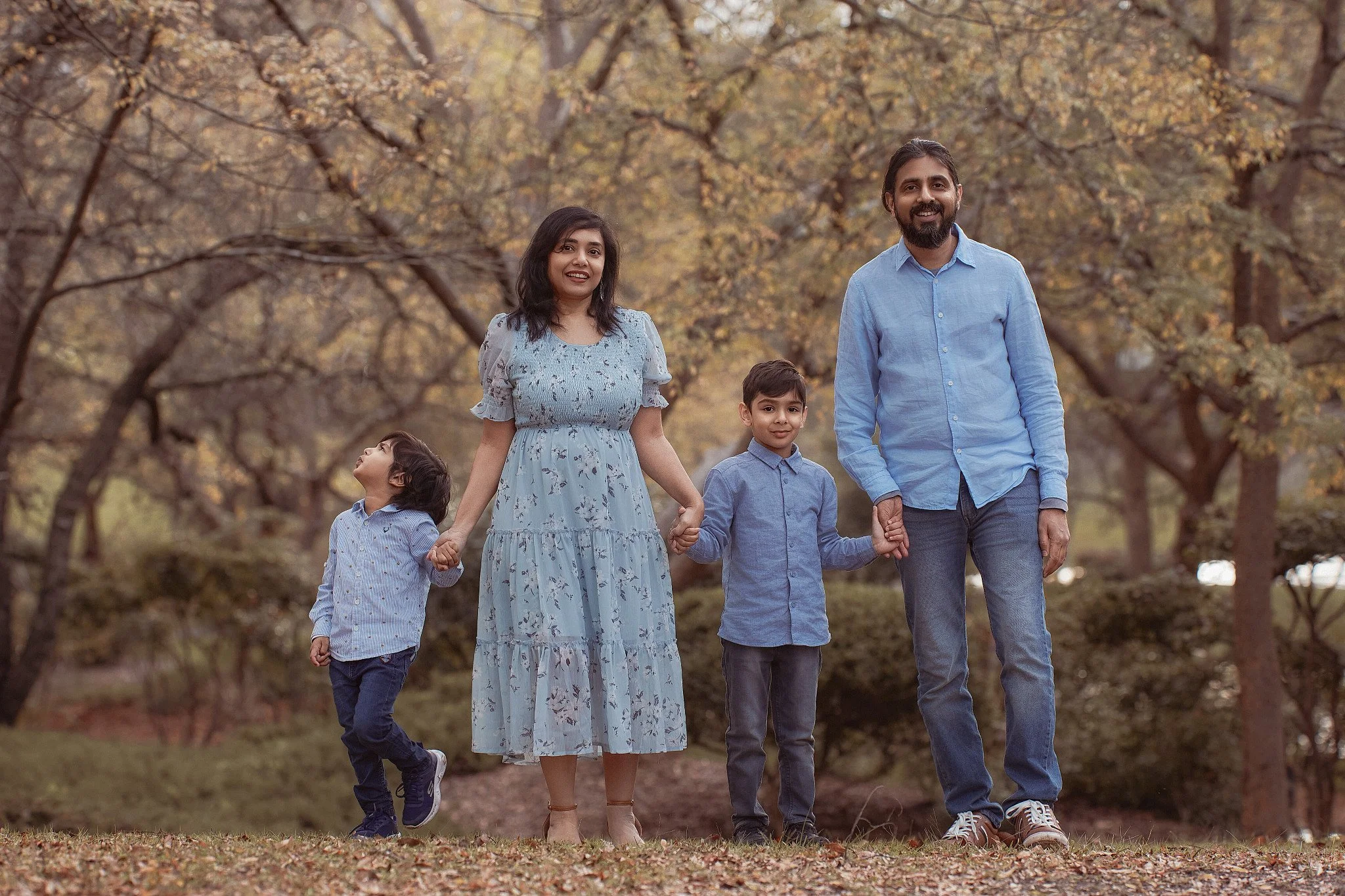 Family walking in park during autumn, holding hands, with trees and fallen leaves in background.