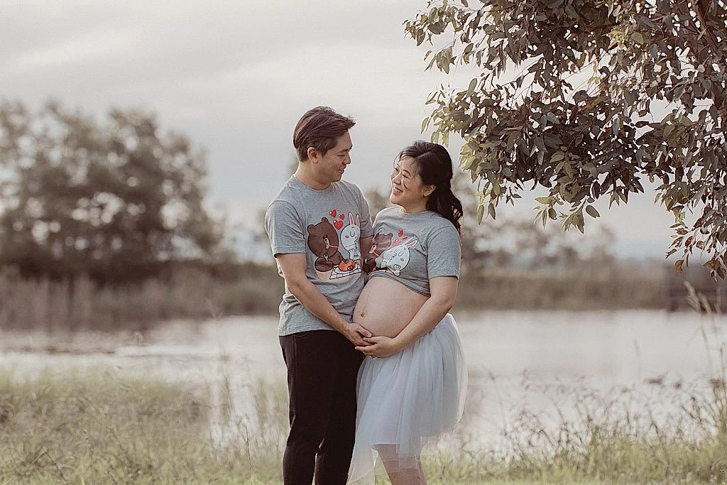 A pregnant woman and a man stand outdoors near a body of water, smiling and holding her belly. They are wearing matching gray t-shirts with cartoon characters.