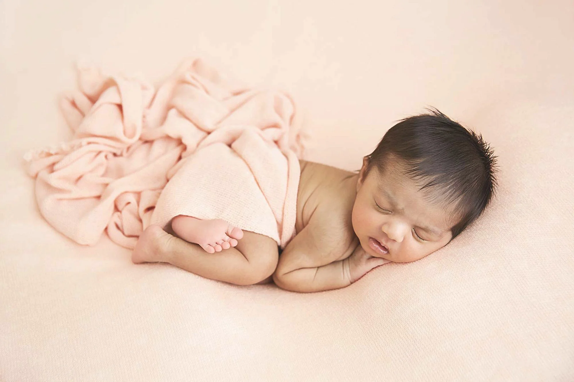 A sleeping baby lying on a soft pink surface, wrapped in a light pink blanket.