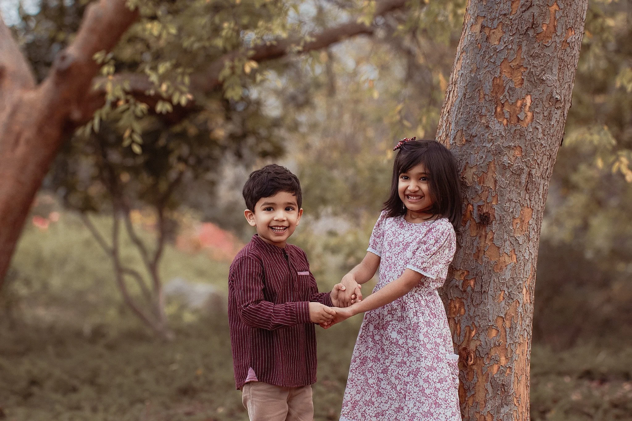 A young boy and girl smiling and holding hands outdoors near a tree in a park, with trees and autumn leaves in the background.