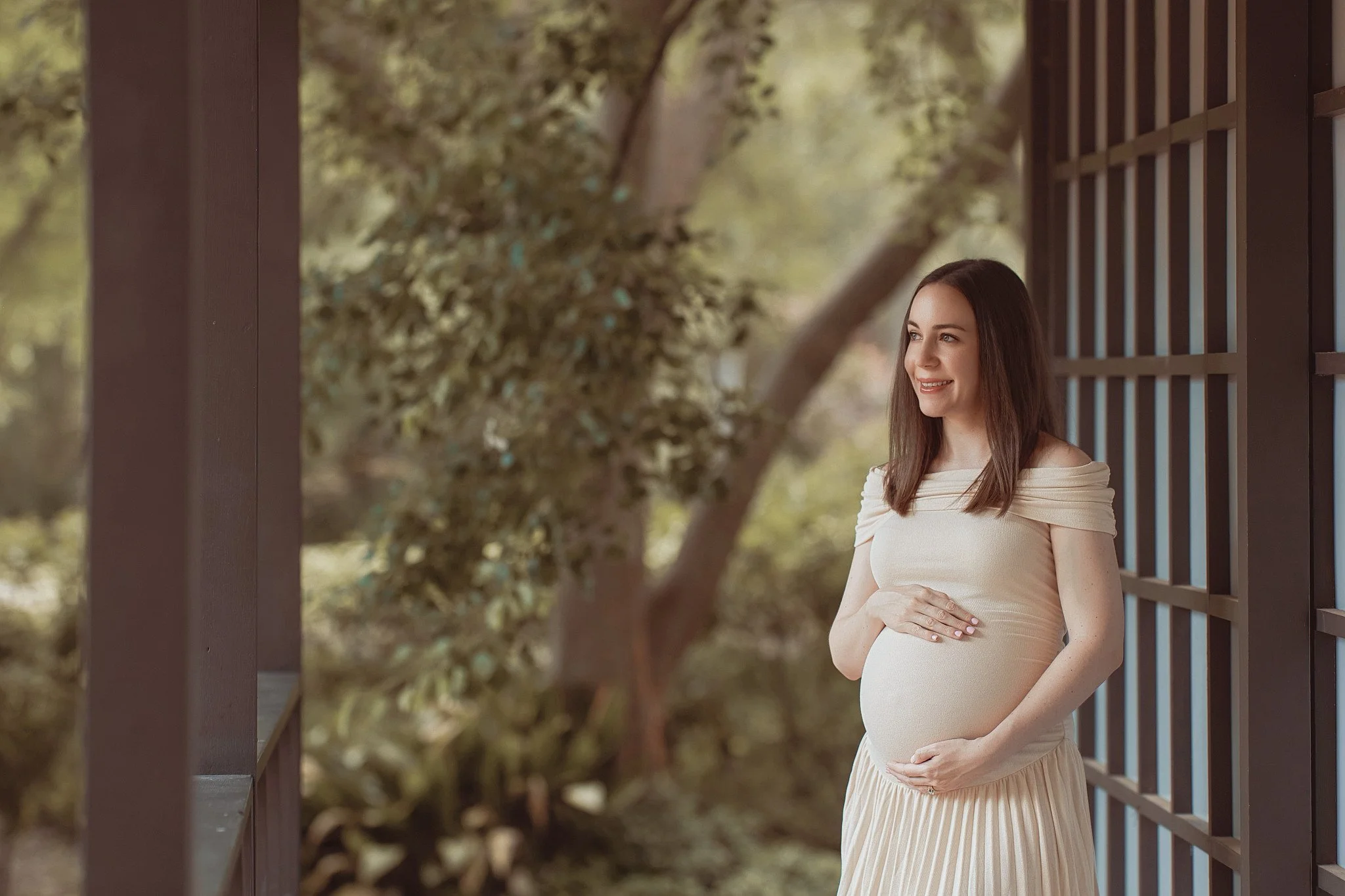 A pregnant woman standing outdoors on a porch, smiling, with her hands resting on her belly, surrounded by greenery.
