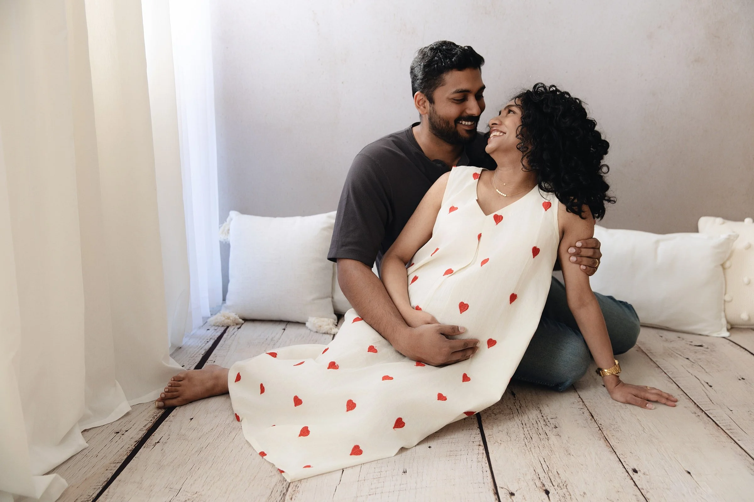 A happy couple sitting on a white rug in a bright room, embracing and smiling at each other. The woman is wearing a white dress with red hearts, and they are both barefoot.