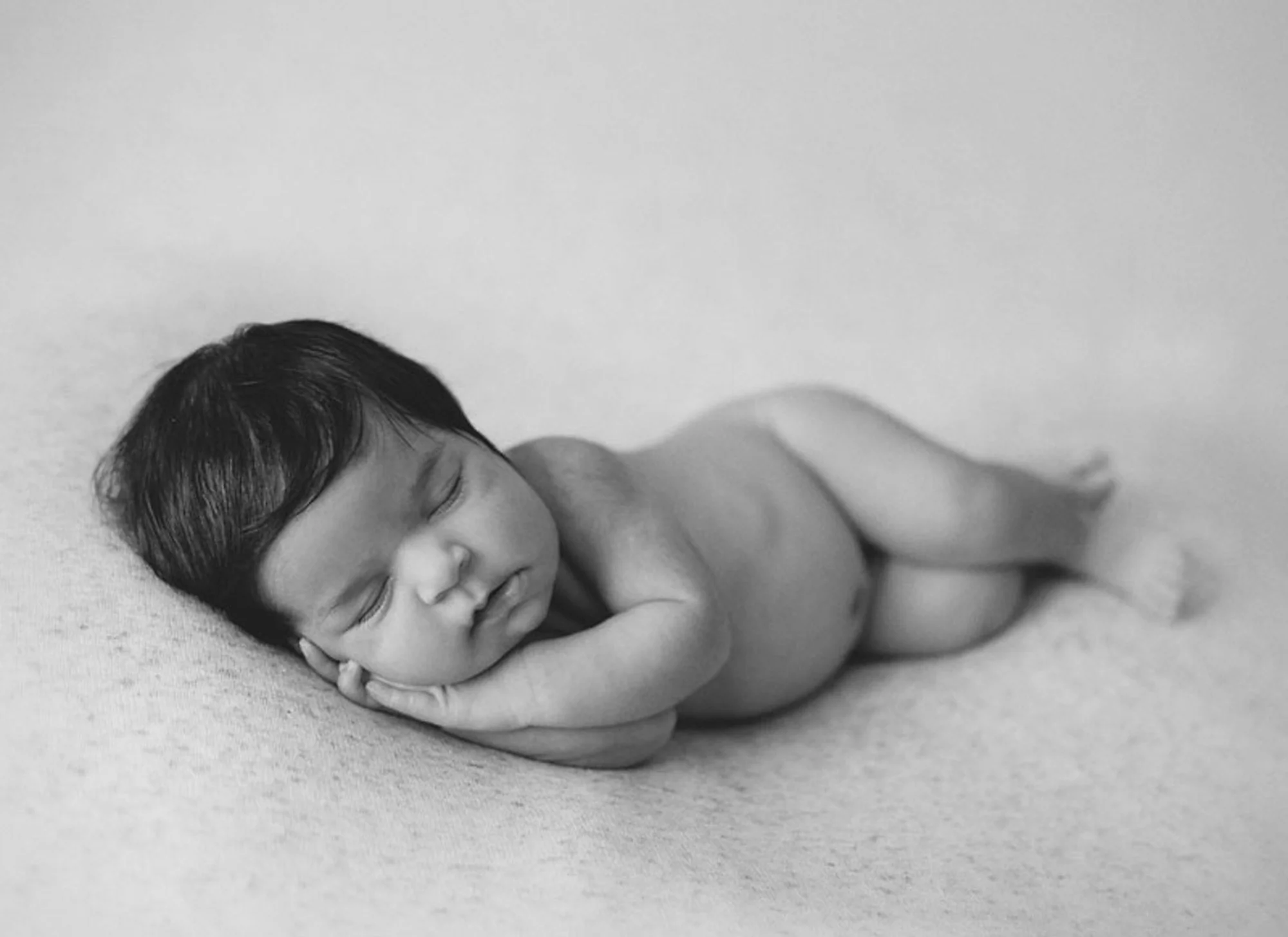 Black and white photo of a newborn baby lying on a soft surface, peacefully sleeping with hands tucked under their chin.