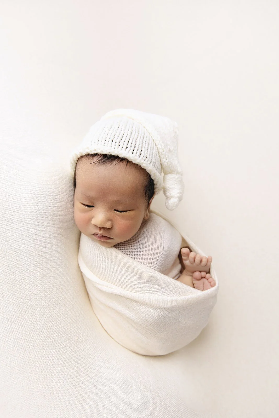 Close-up of a young Asian baby swaddled in cream-colored fabric, wearing a white knit hat with a pom-pom, lying on a matching cream background.