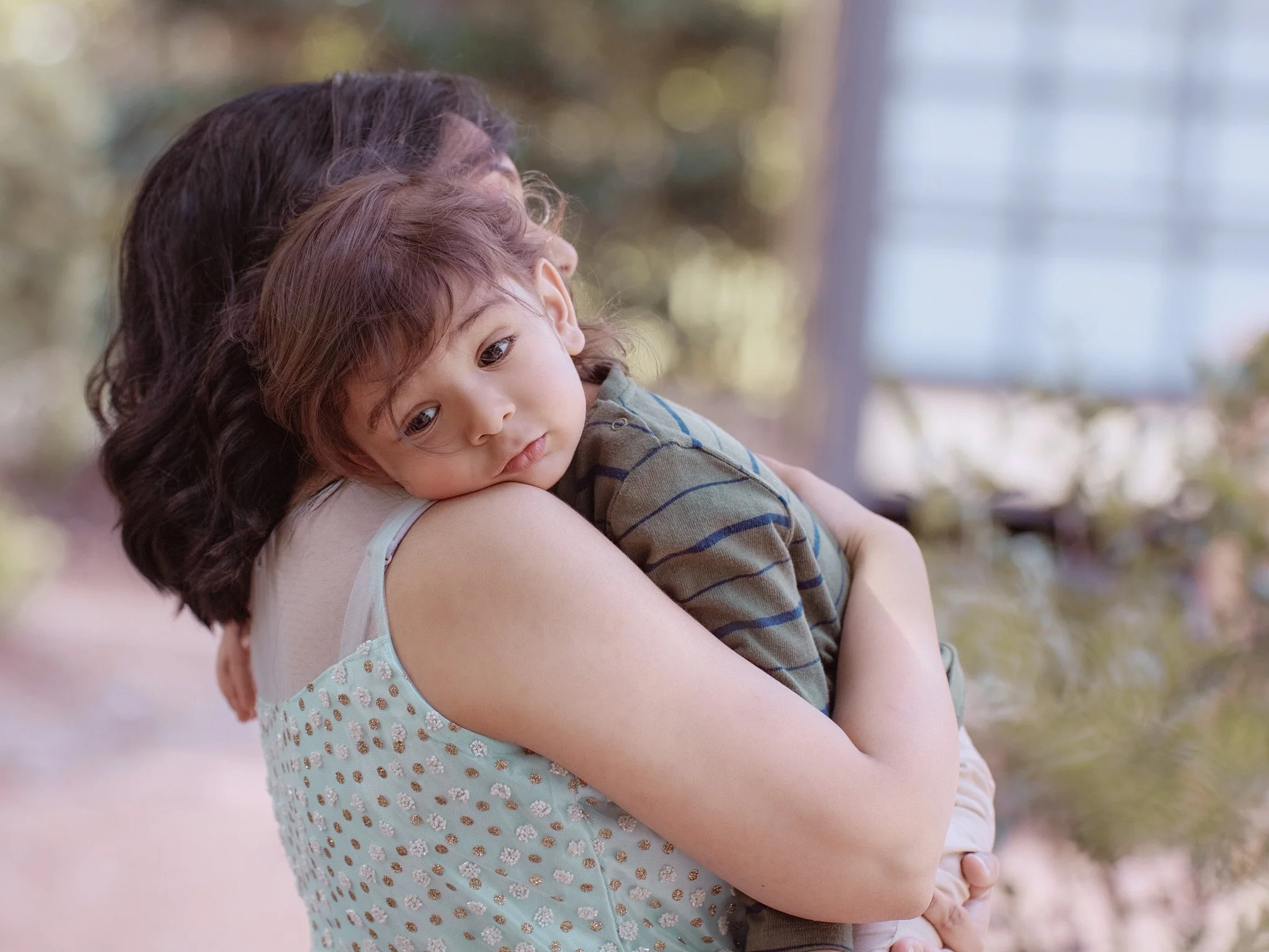 A woman holding a young boy close, both with calm expressions, outdoors with blurred trees and a building in the background.
