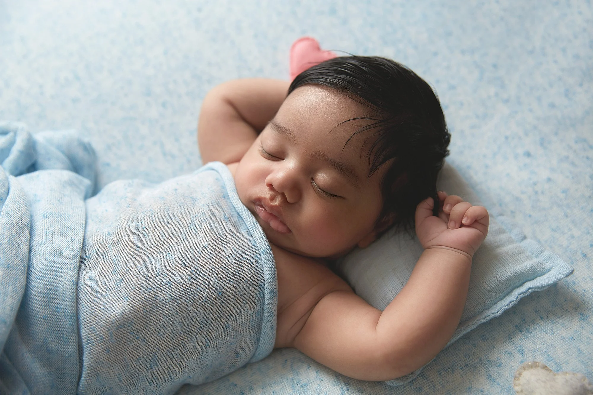 A sleeping baby with dark hair, lying on a light blue blanket with a pillow, resting peacefully with arms bent and one hand near the ear.