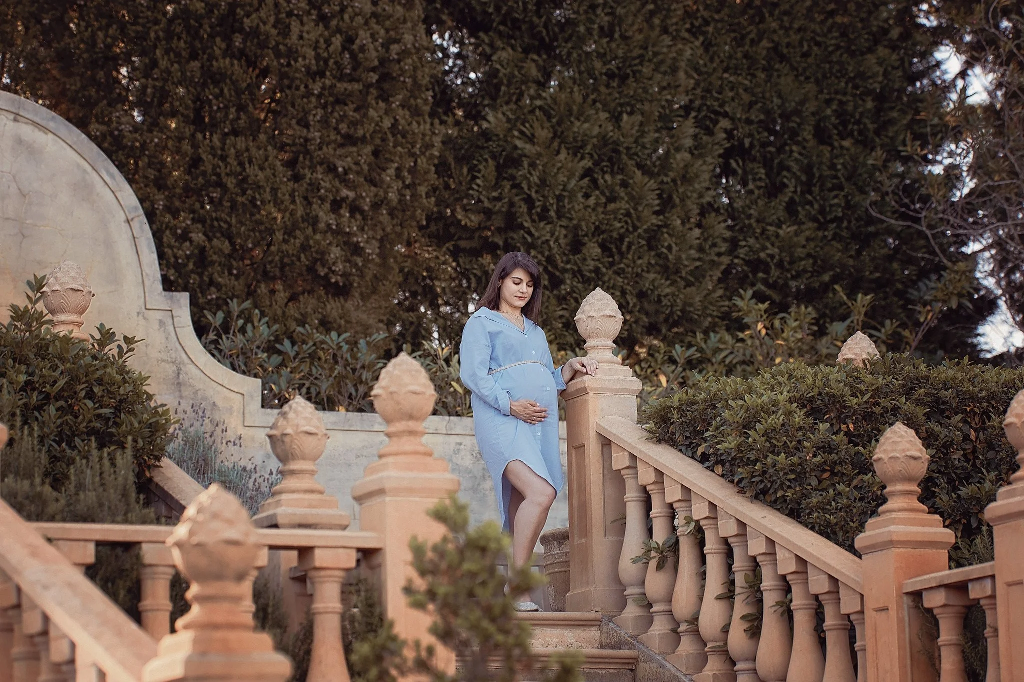 Pregnant woman in blue dress standing on outdoor staircase surrounded by bushes and trees, gazing downward with hand on her belly.