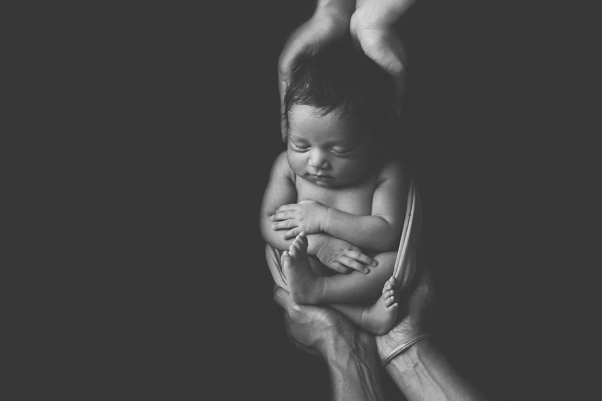 Black and white photo of a newborn baby cradled gently in an adult's hands, with the hands supporting the baby's head and body, looking peaceful and content.