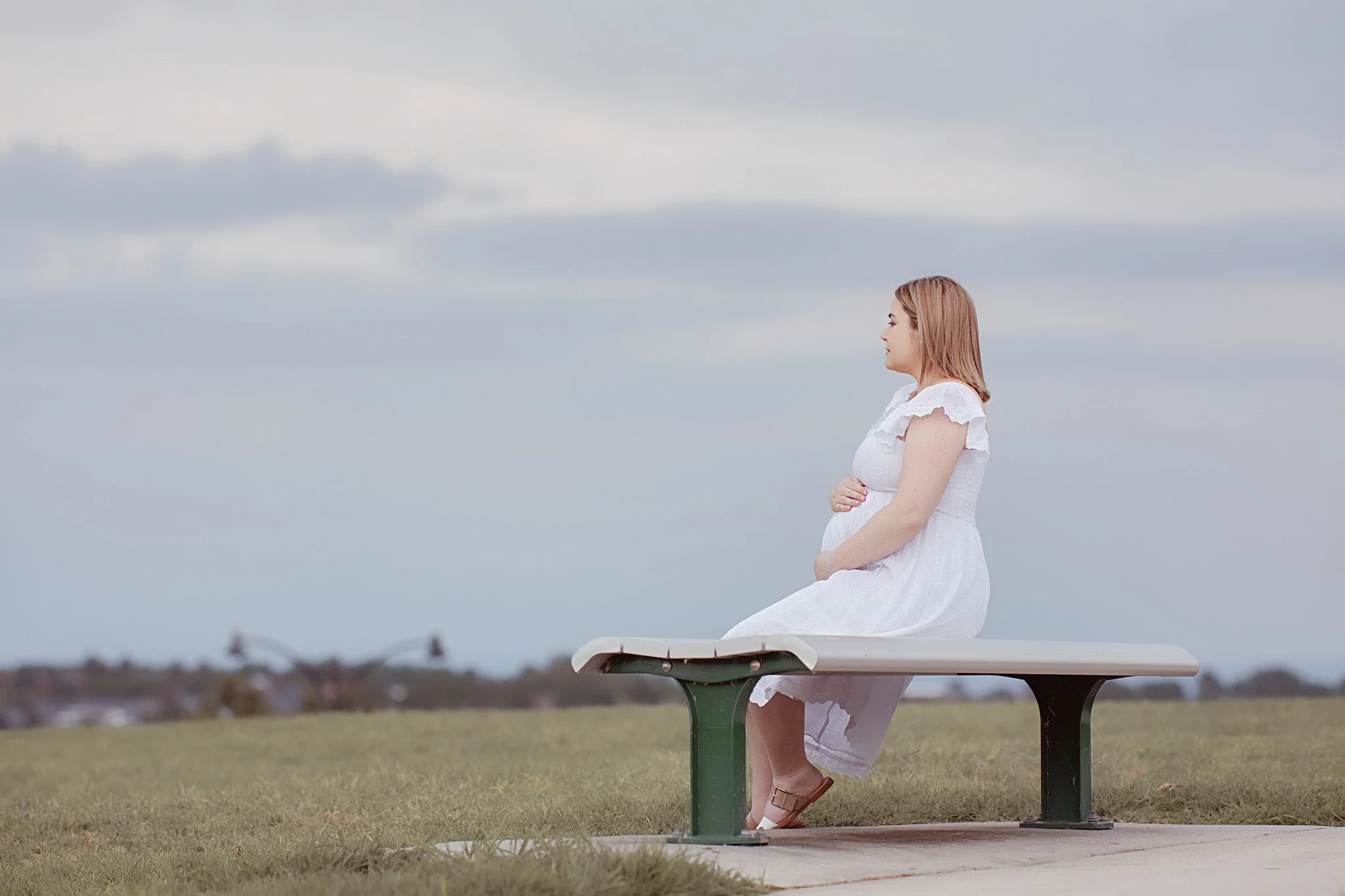 A pregnant woman sitting on a park bench, looking to the side with arms crossed over her belly, wearing a white dress, with a cloudy sky and open grassy field in the background.