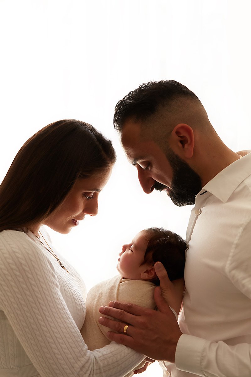 A couple holds their newborn baby, with their foreheads touching, inside a softly lit room.
