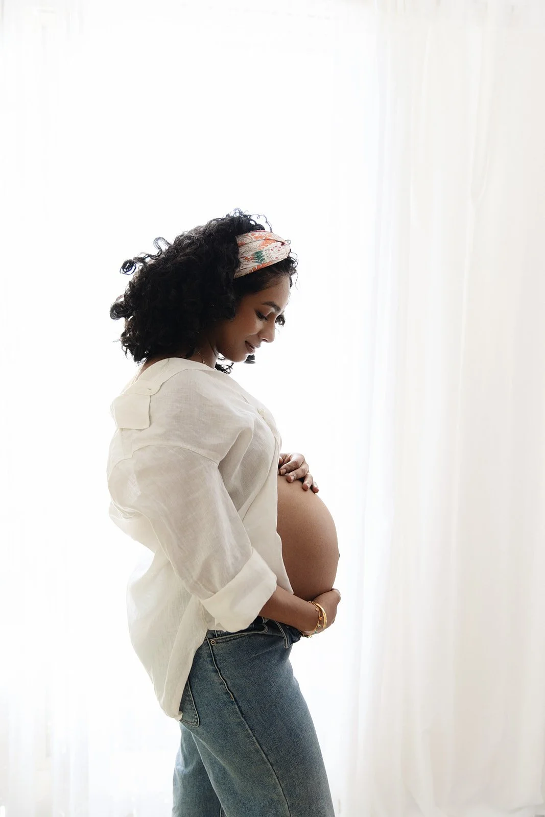 Pregnant woman in white blouse and jeans standing near a bright window, holding her belly and smiling.