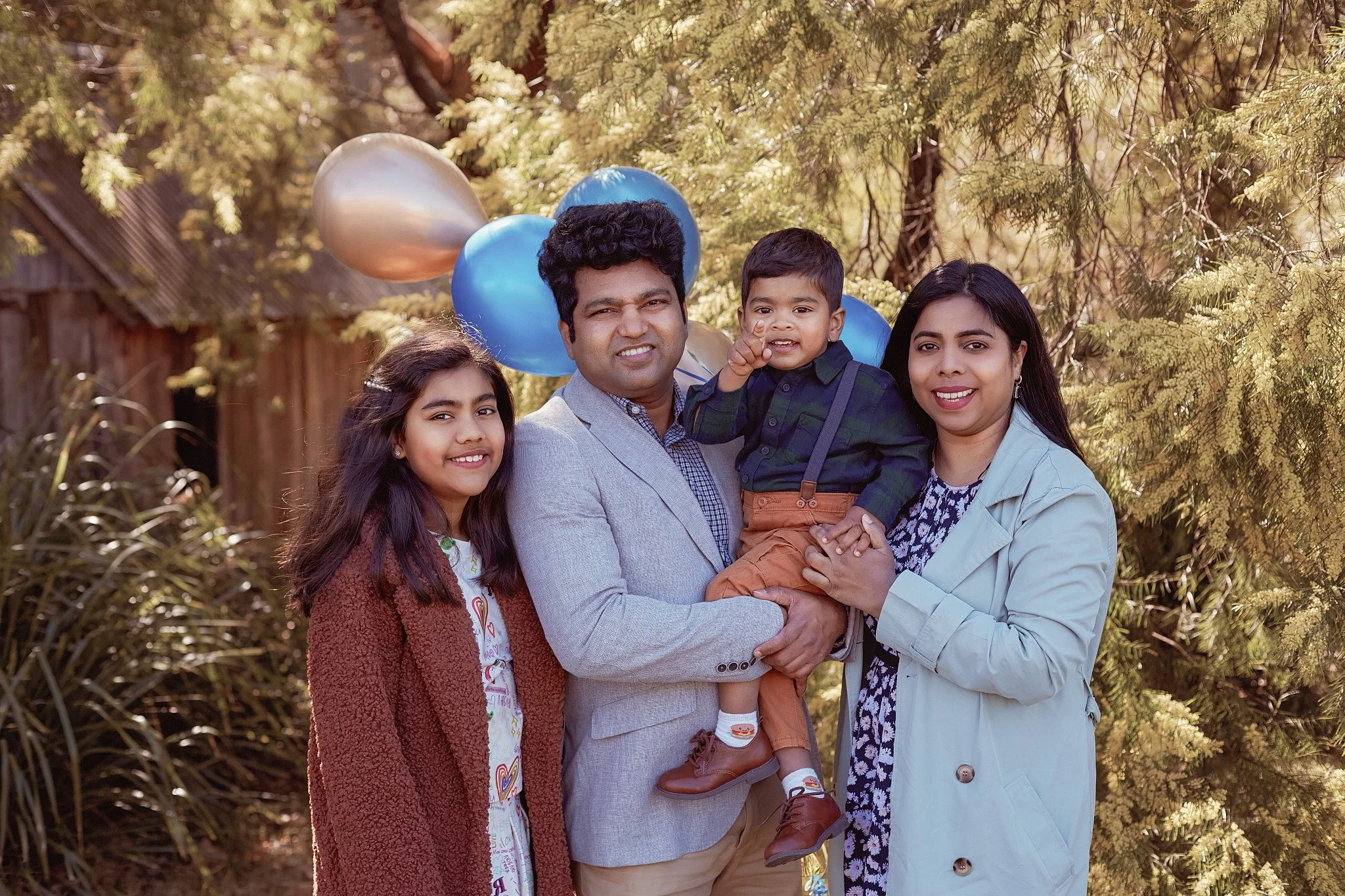 A family of four posing outdoors with balloons in the background, surrounded by trees and greenery, smiling at the camera.