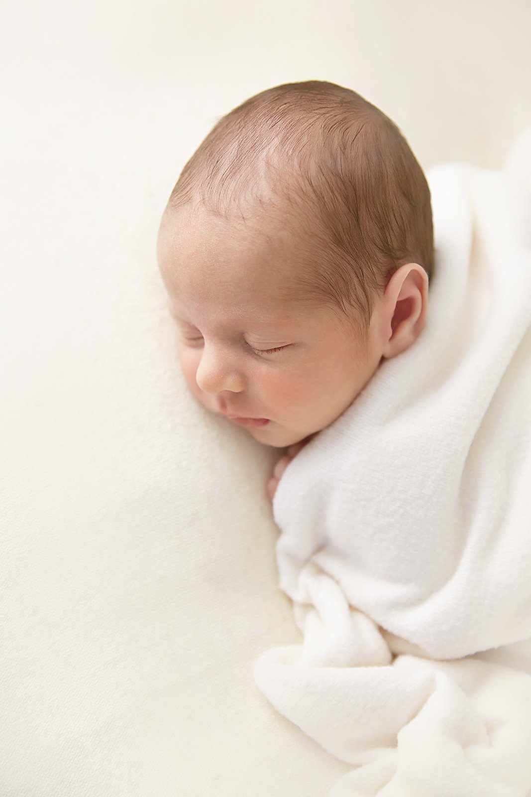 Close-up of a sleeping newborn baby with light brown hair, wrapped in a white blanket, lying on a soft cream-colored surface.