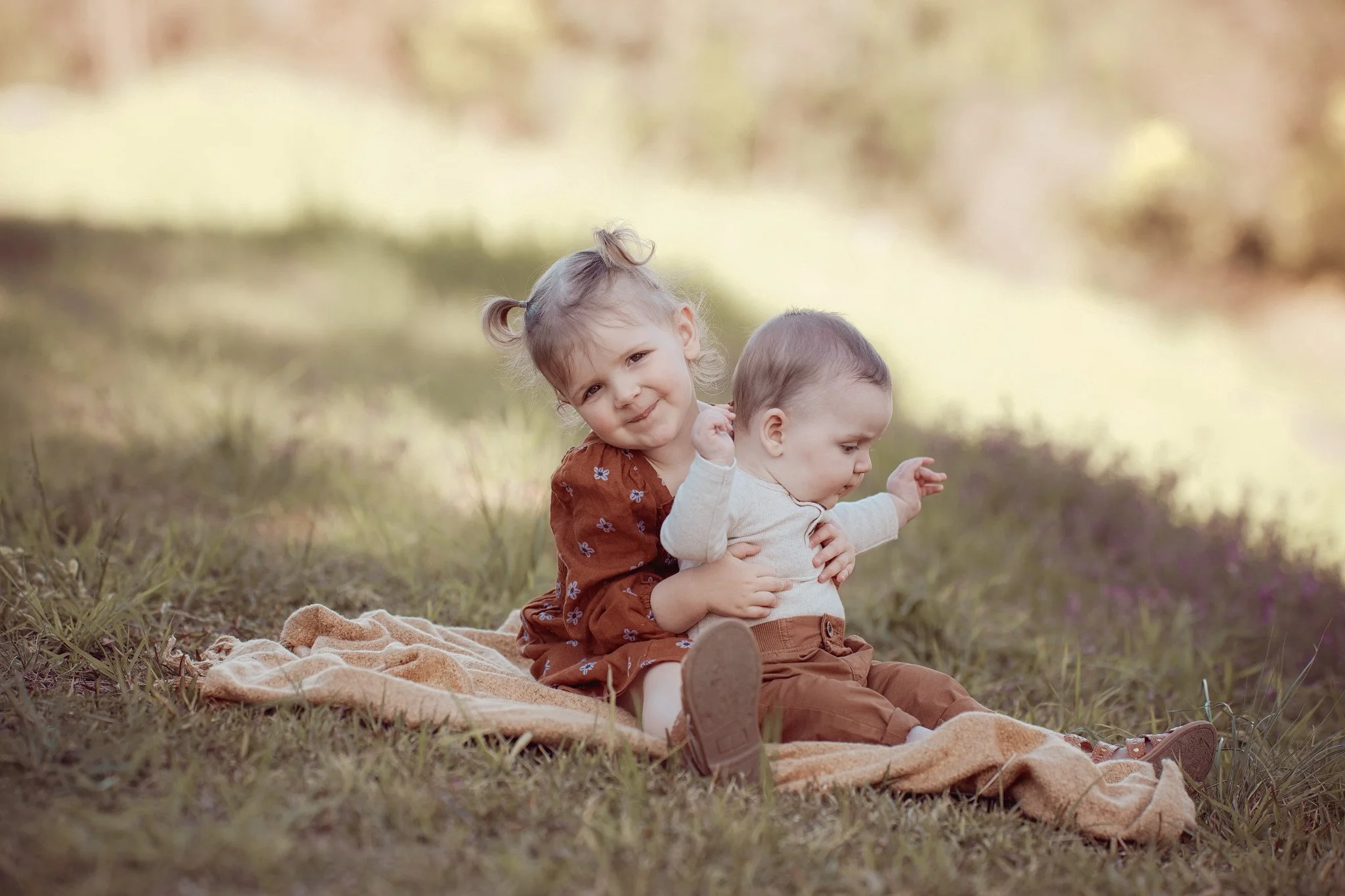 A young girl sitting on a blanket outdoors, hugging a baby boy, both dressed in brown clothing, on a grassy field with blurred background.