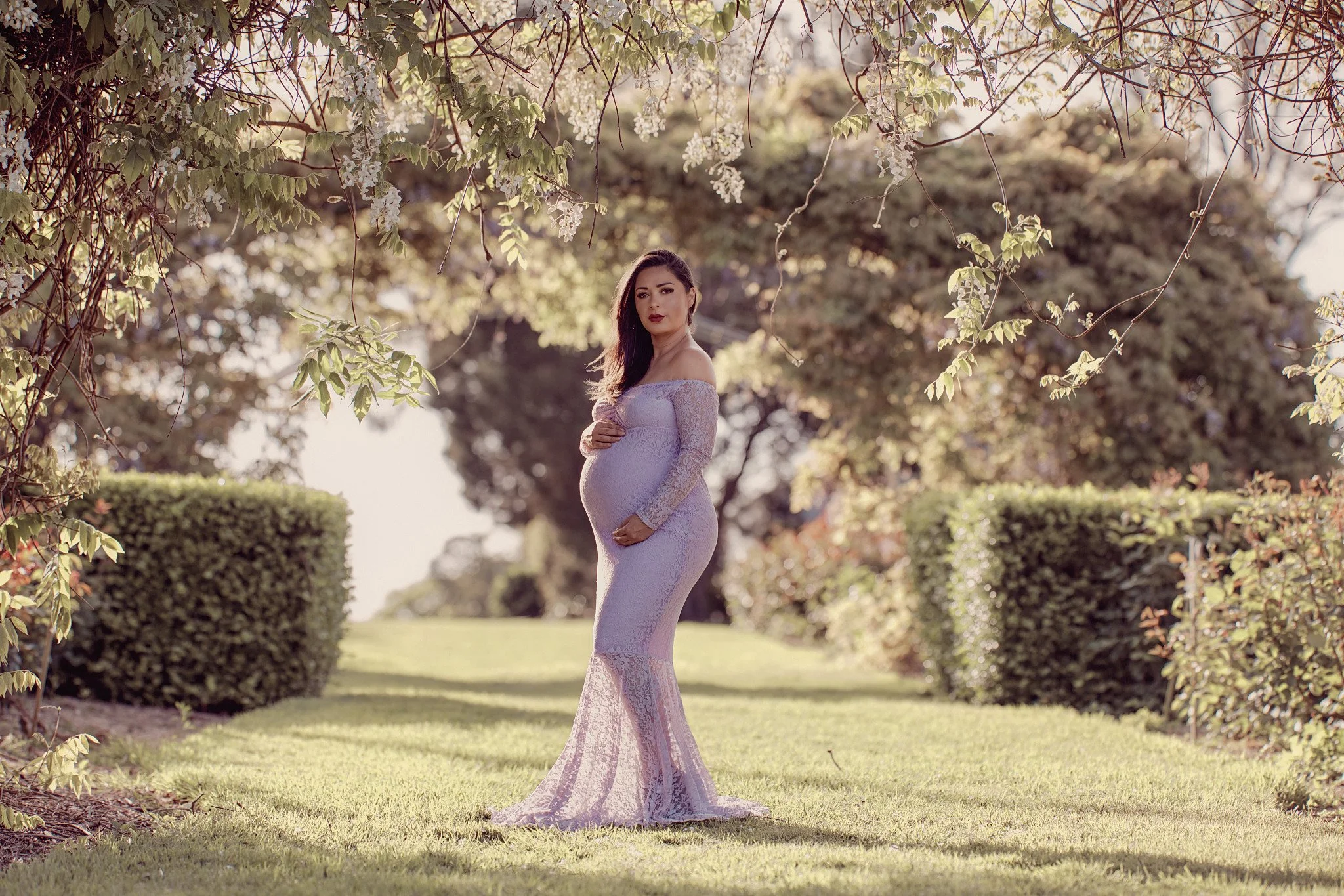 A pregnant woman in a light purple lace dress standing on a grassy pathway under trees and bushes during daytime.