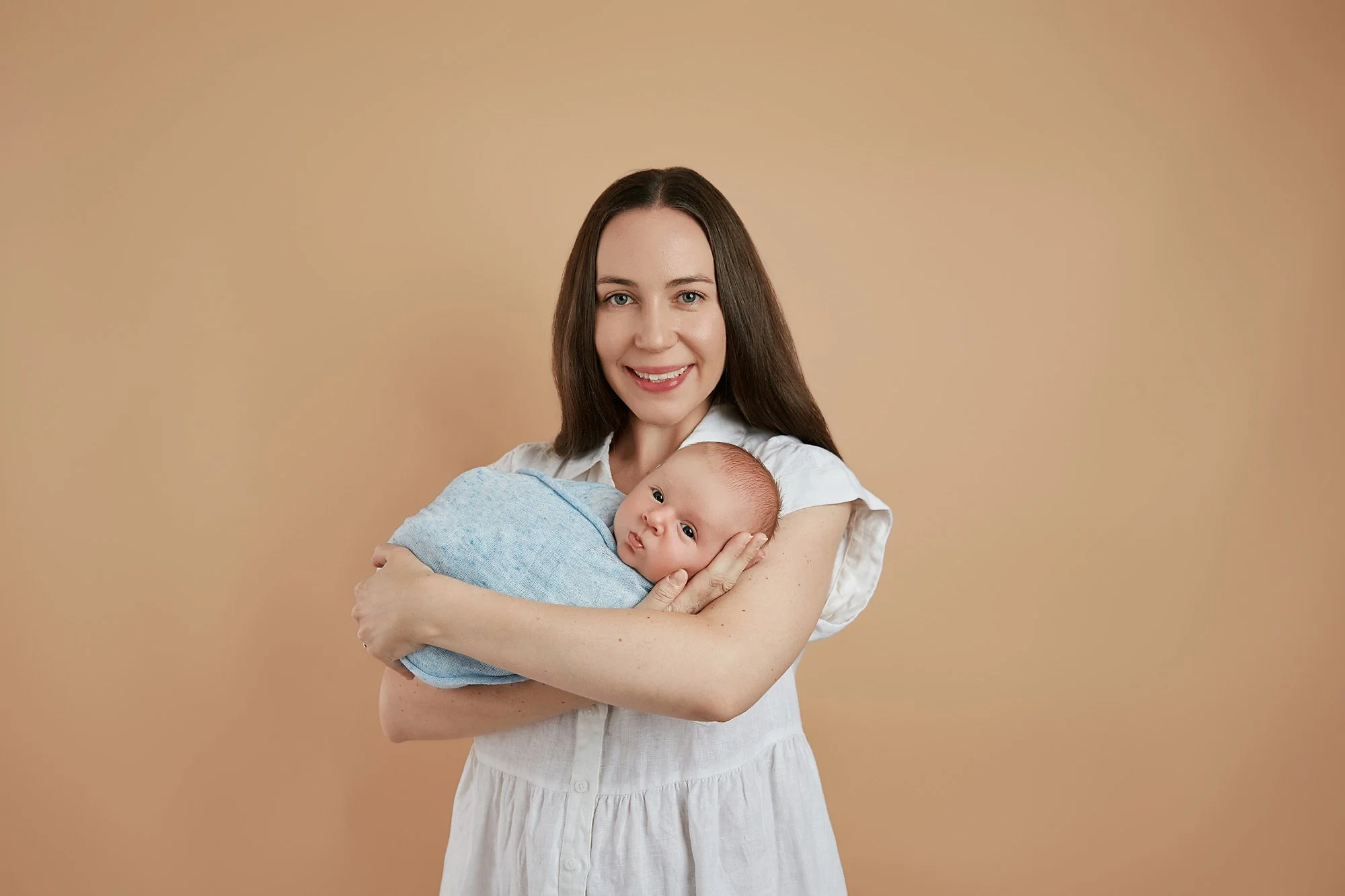 A woman with long brown hair holding a baby wrapped in a blue blanket against a beige background.