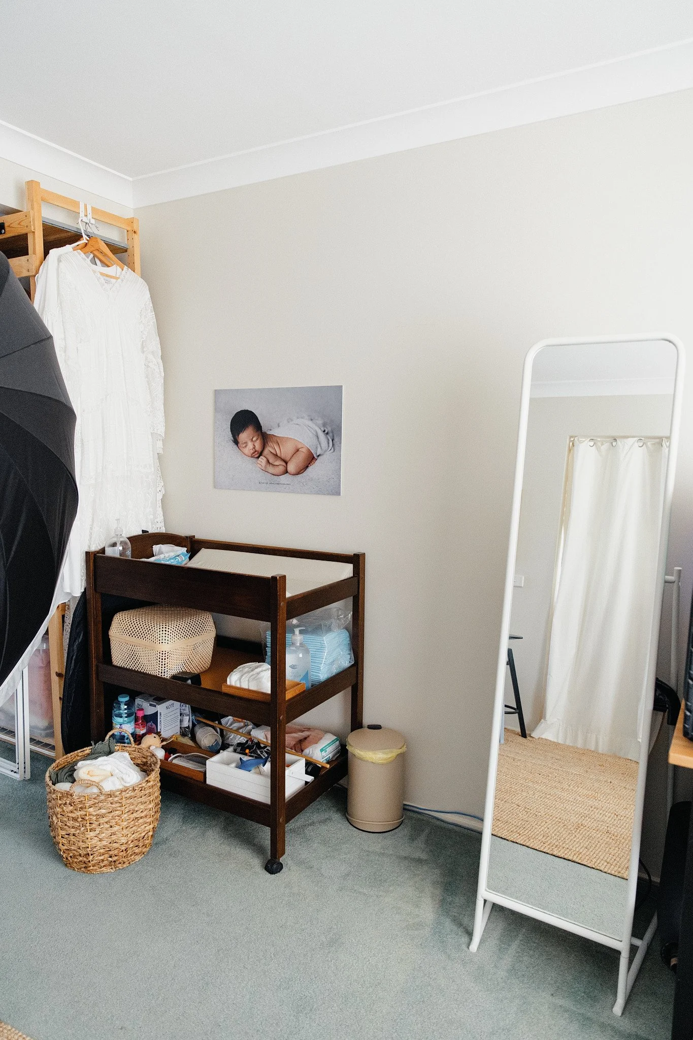 A newborn studio with a baby changing station, a clothing rack, a mirror, and a posed baby portrait of a sleeping infant on the wall.