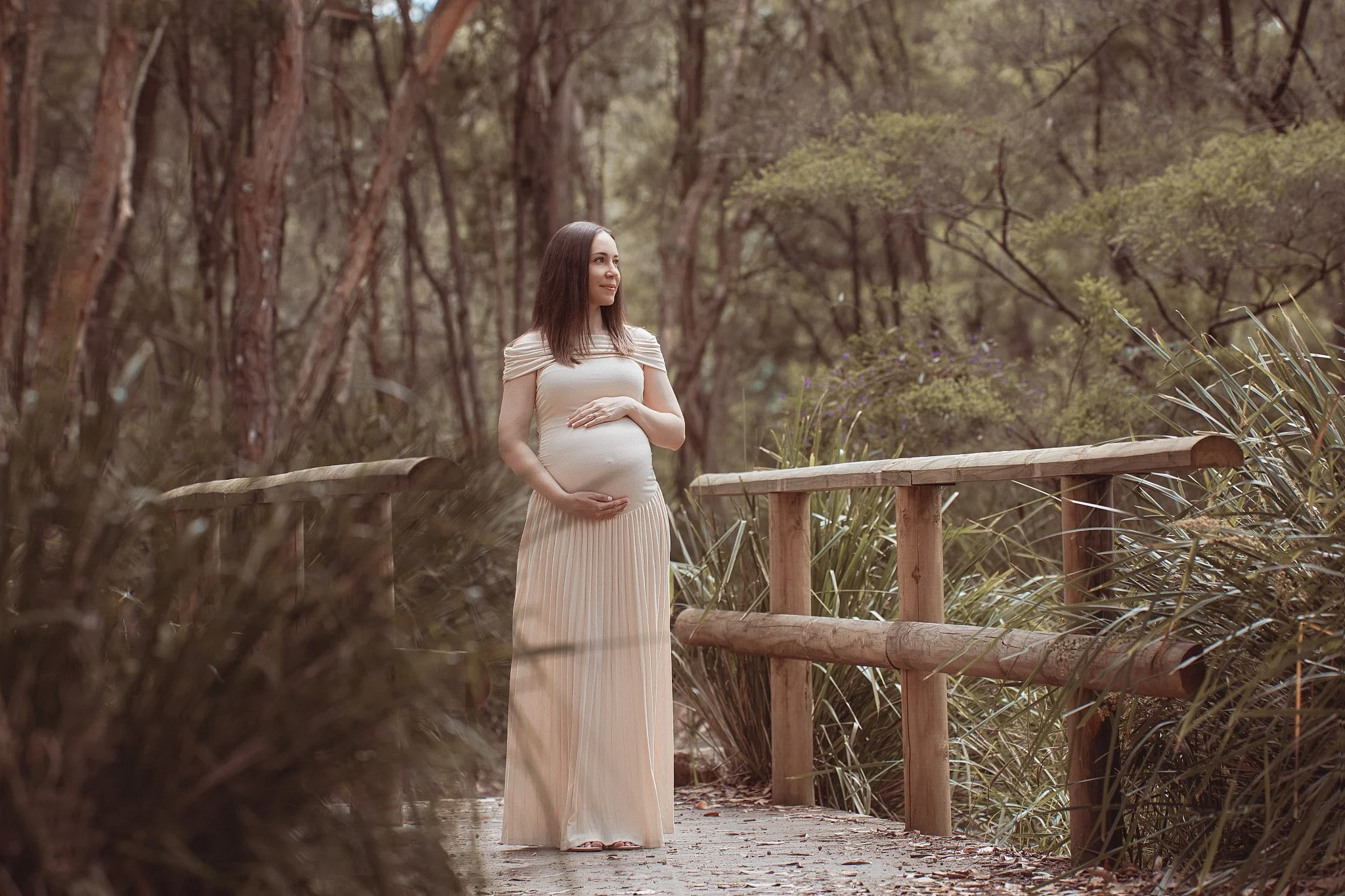 A pregnant woman standing on a wooden path in a forest, wearing a white dress, with her hands on her belly, looking to the side.