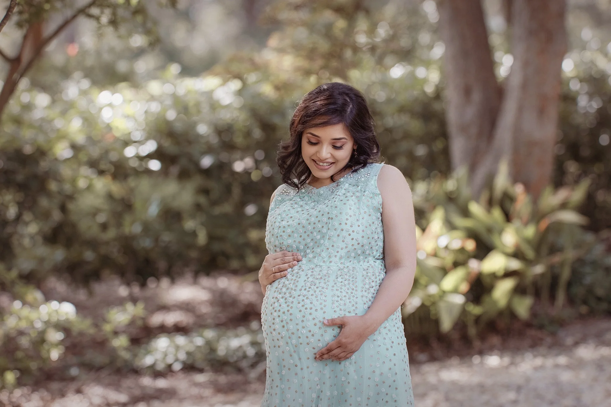 A pregnant woman in a light blue dress with small patterns, standing outdoors in a natural setting with trees and foliage, looking down at her belly with a gentle smile, cradling her stomach with both hands.