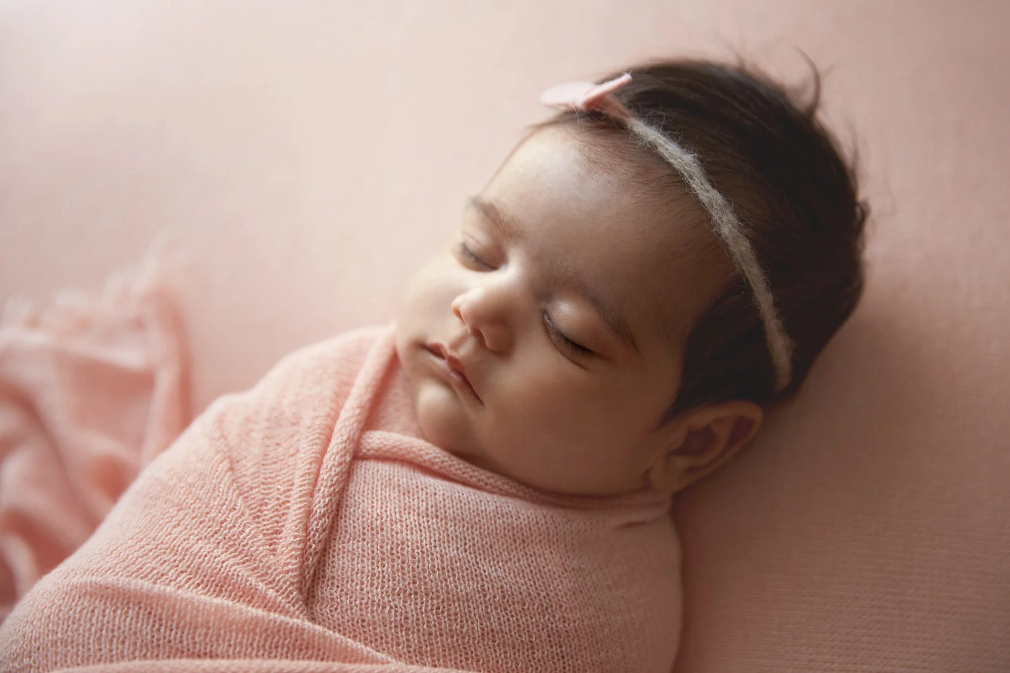 A young girl with dark hair, peacefully sleeping, wrapped in a pink blanket, lying on a soft pink surface.