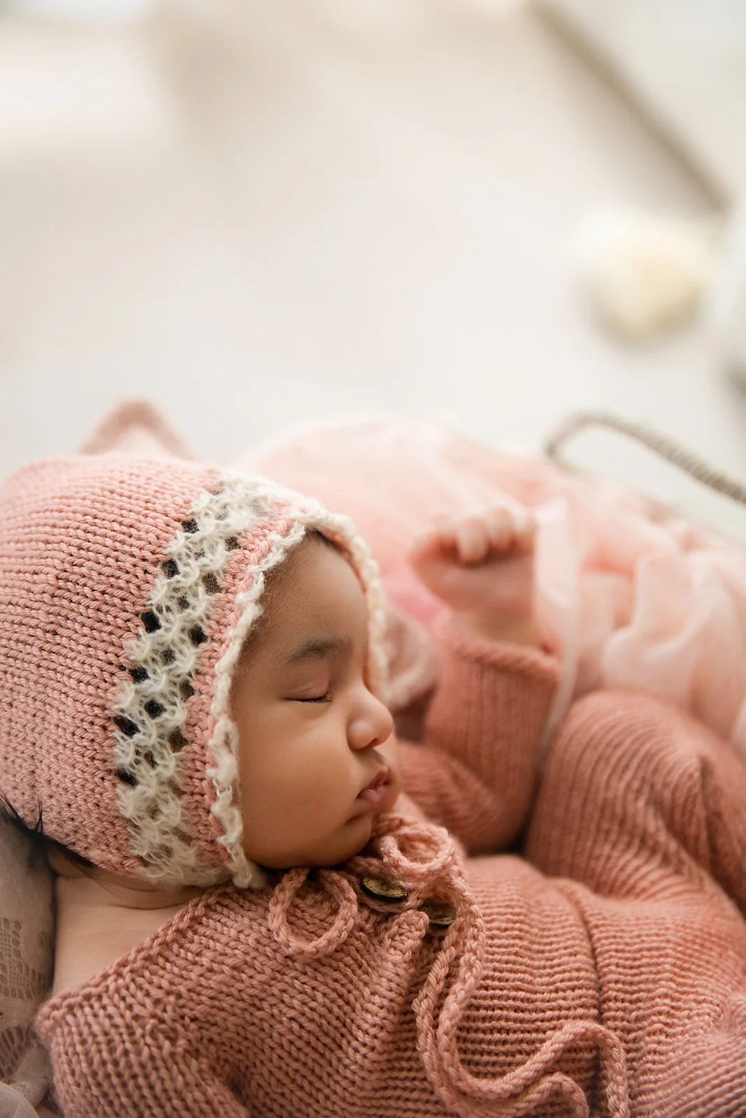 A young child with closed eyes sleeping, wearing a pink knit hat and matching pink sweater, resting with their head on a soft surface.