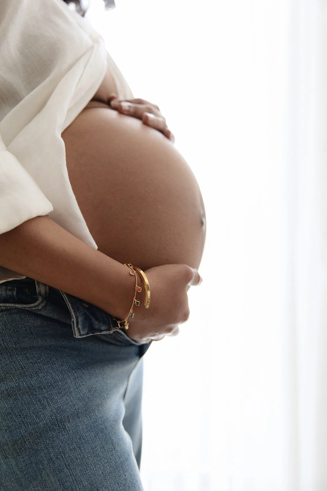 Close-up of a pregnant woman holding her belly, wearing a white shirt, dark jeans, and gold jewelry