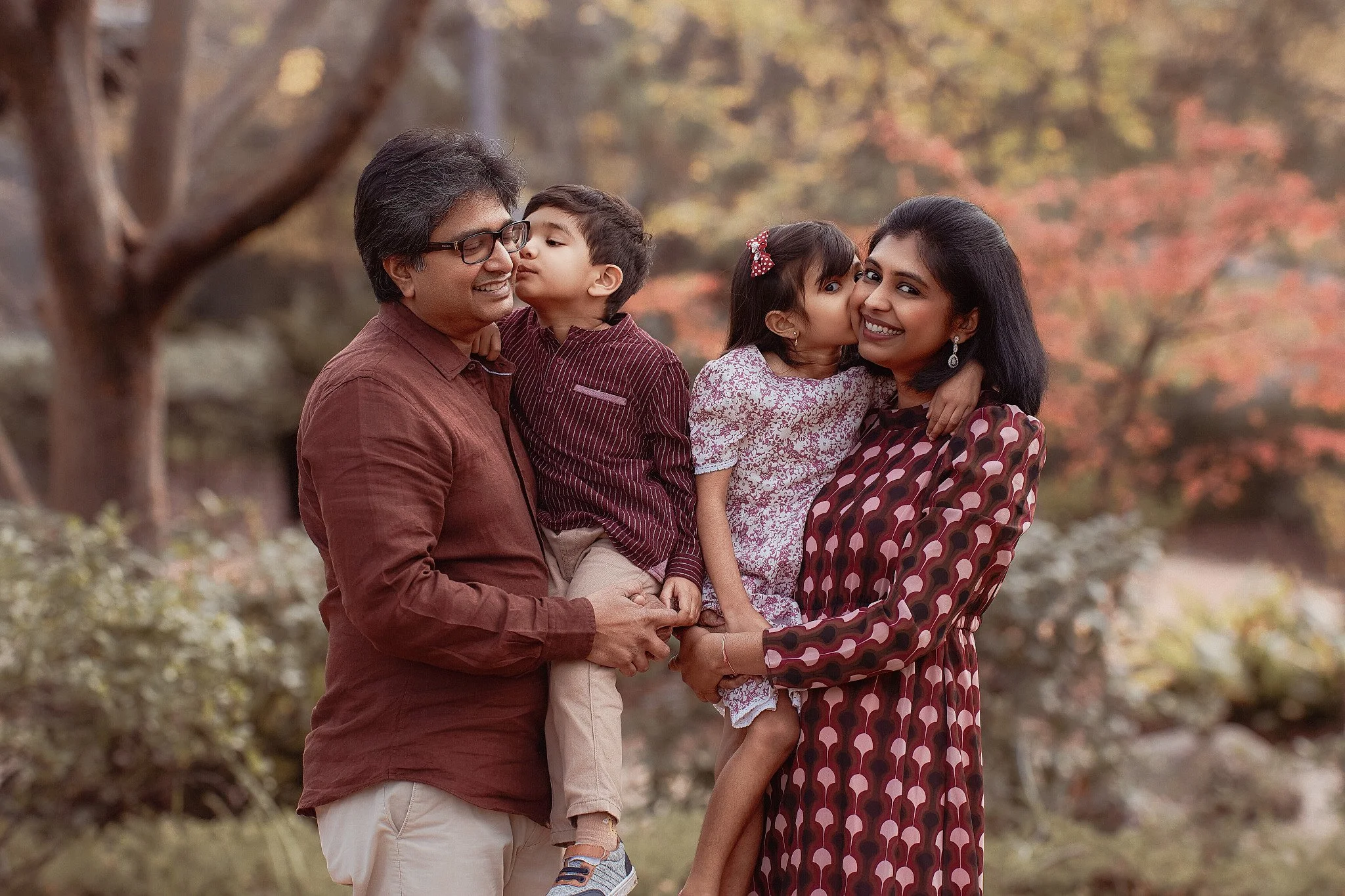 A smiling family of four outdoors in autumn, with trees in fall colors in the background. The father holds one son, and the mother holds a daughter, who kisses the mother on the cheek.