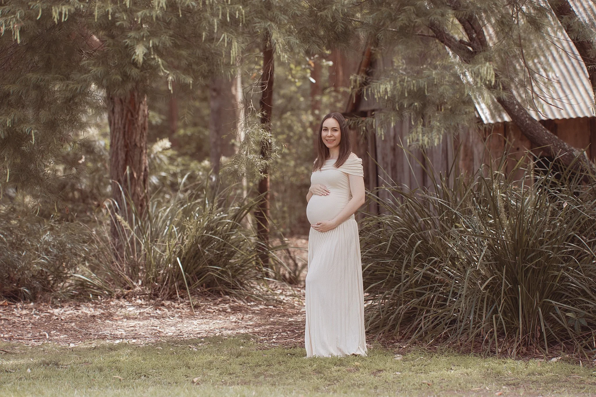 A pregnant woman standing outdoors in front of trees and a rustic wooden building, wearing a long white dress and smiling.