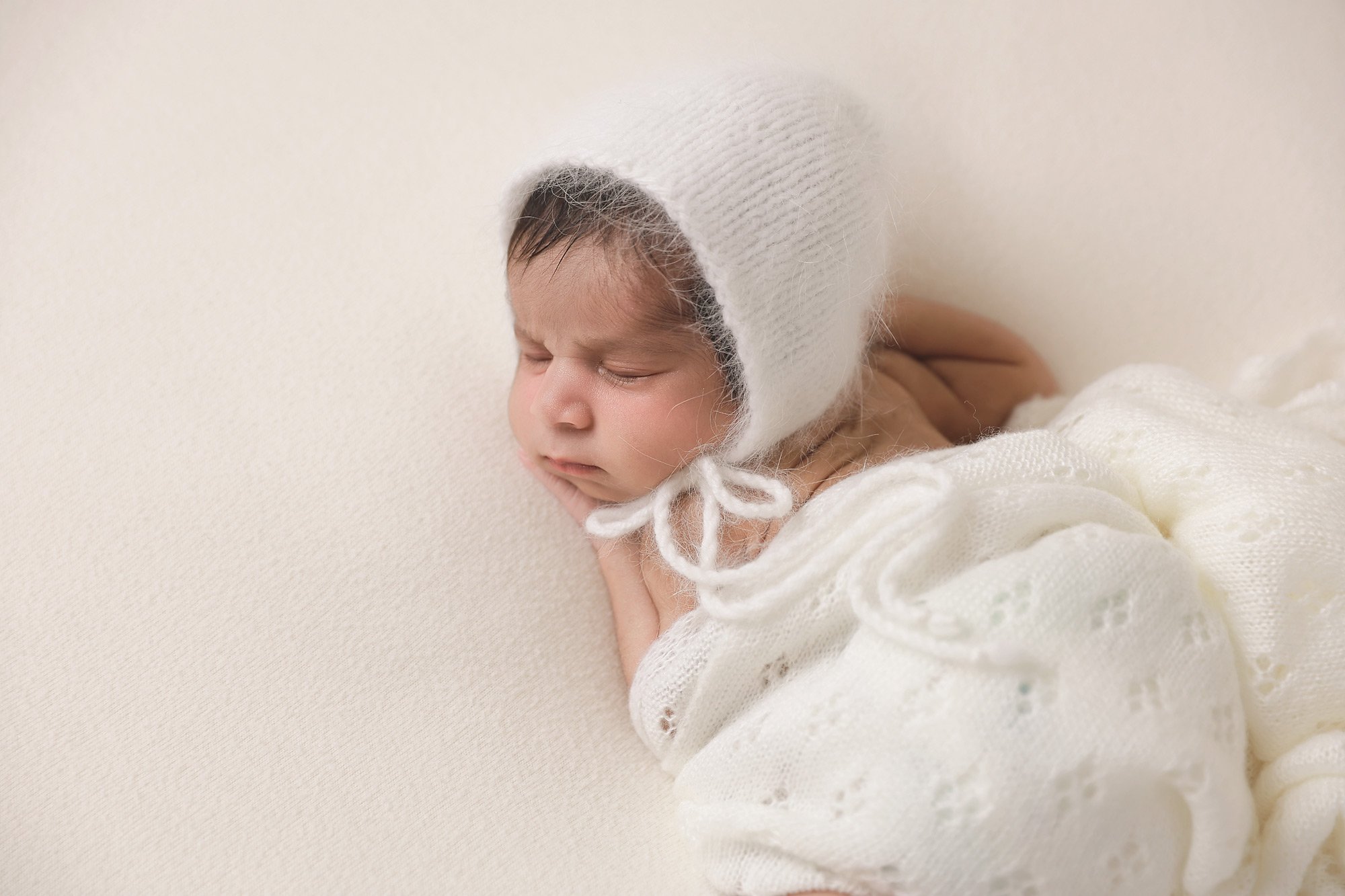 A newborn baby wearing a knitted white bonnet and a white lace blanket, sleeping on a cream-colored surface.