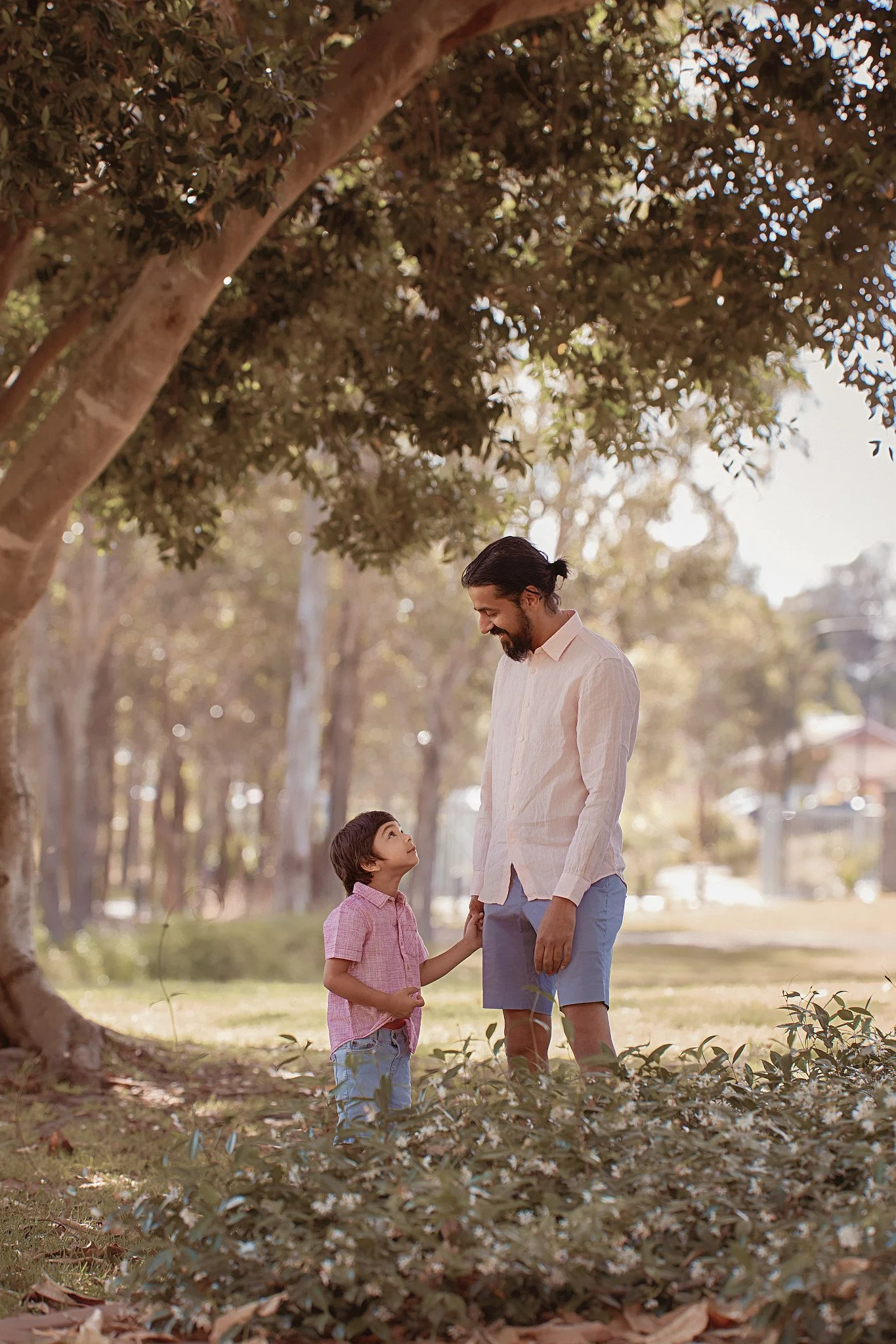 A man and a young boy standing outdoors under a large tree, smiling at each other, with greenery and a park-like setting in the background.