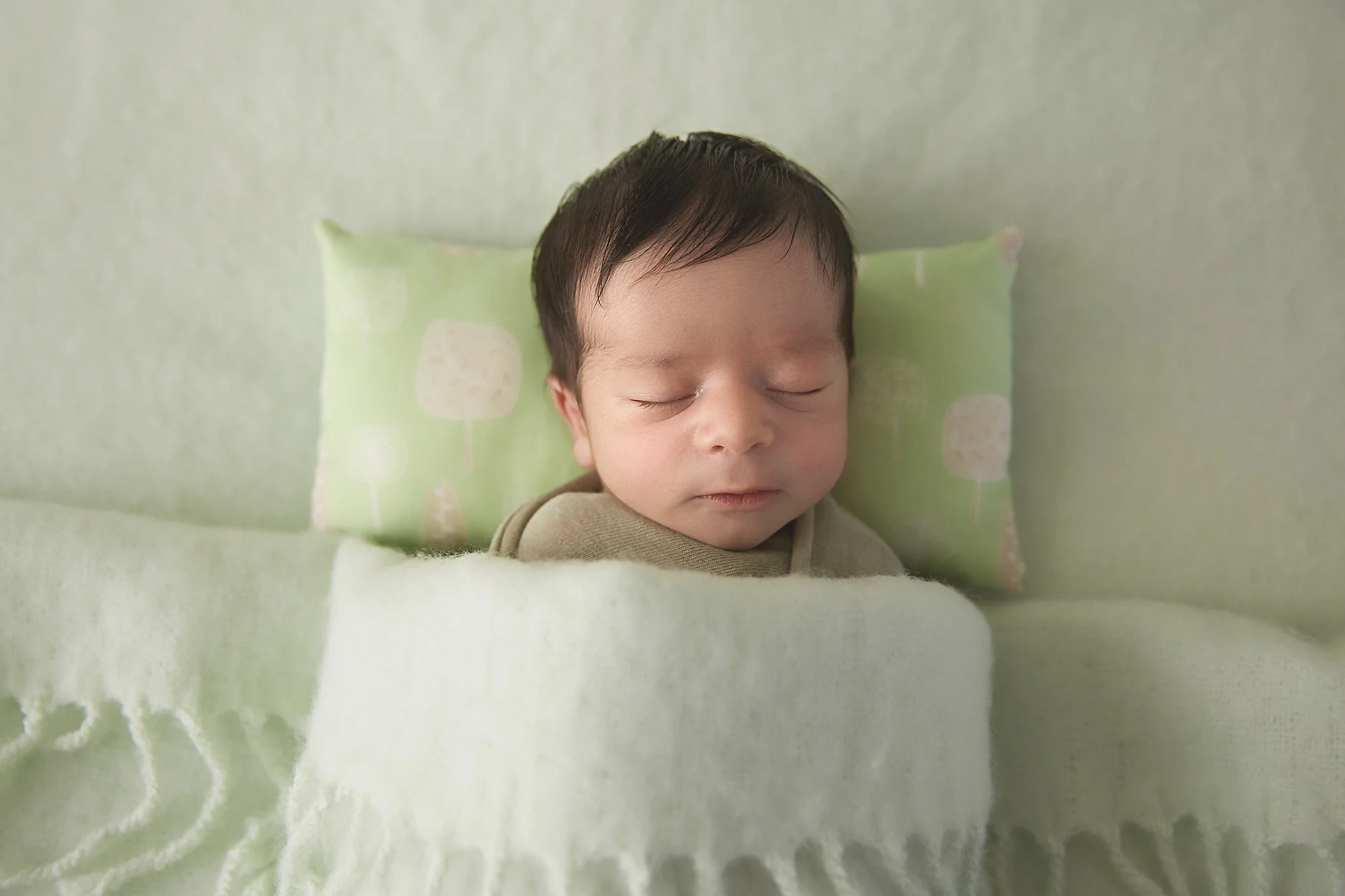 A young child with dark hair sleeping peacefully on a green pillow with a white, soft blanket covering them in a cozy setting.