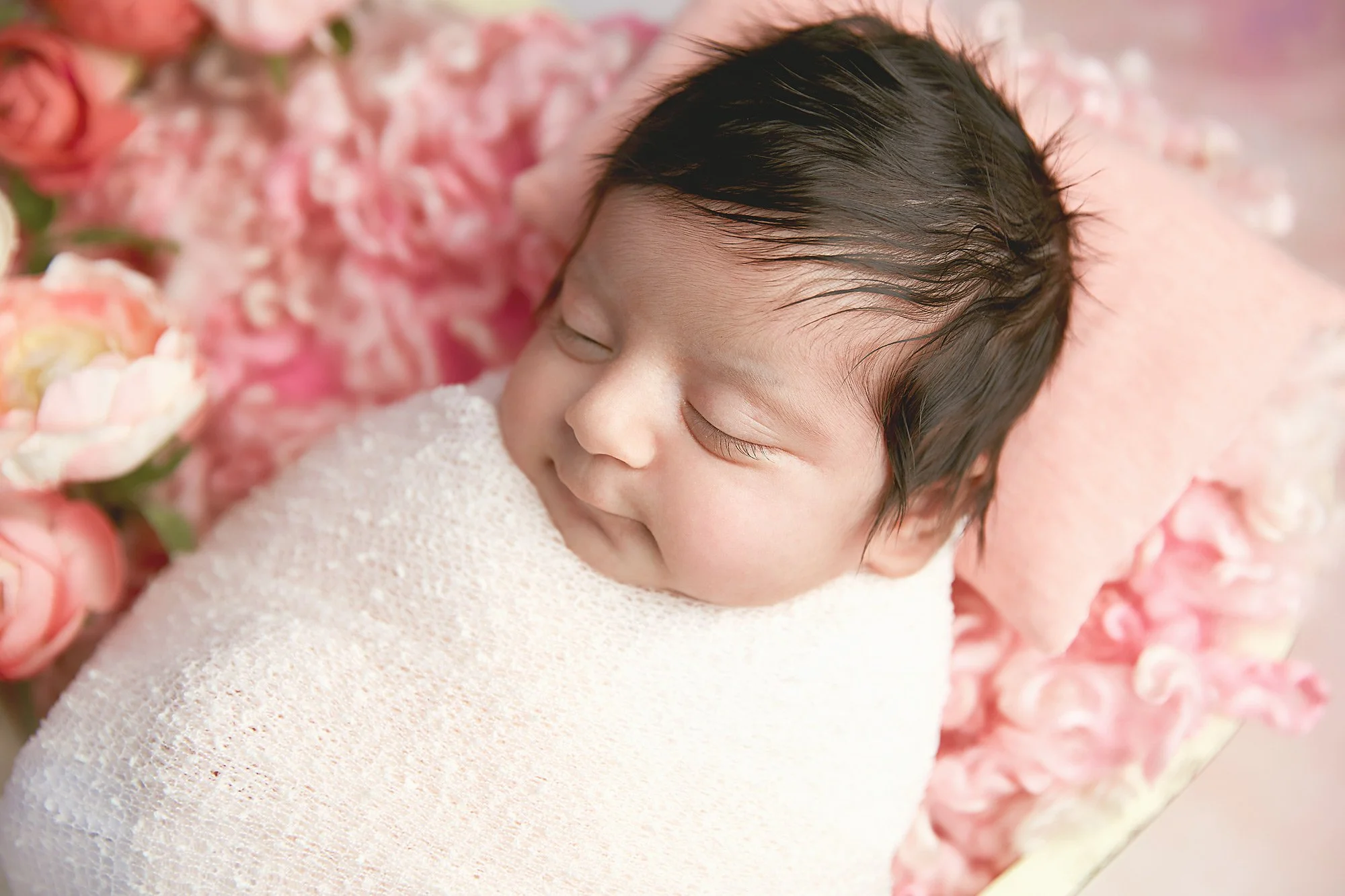 Close-up of a sleeping newborn baby wrapped in a white blanket, lying on a pink and floral background.