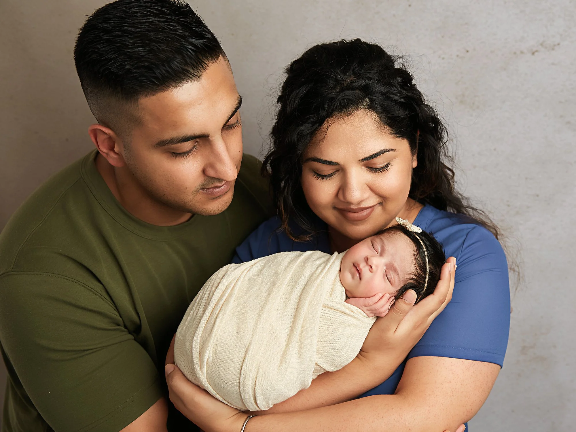 A couple holding a sleeping newborn baby wrapped in a beige blanket, all with closed eyes and smiling softly.