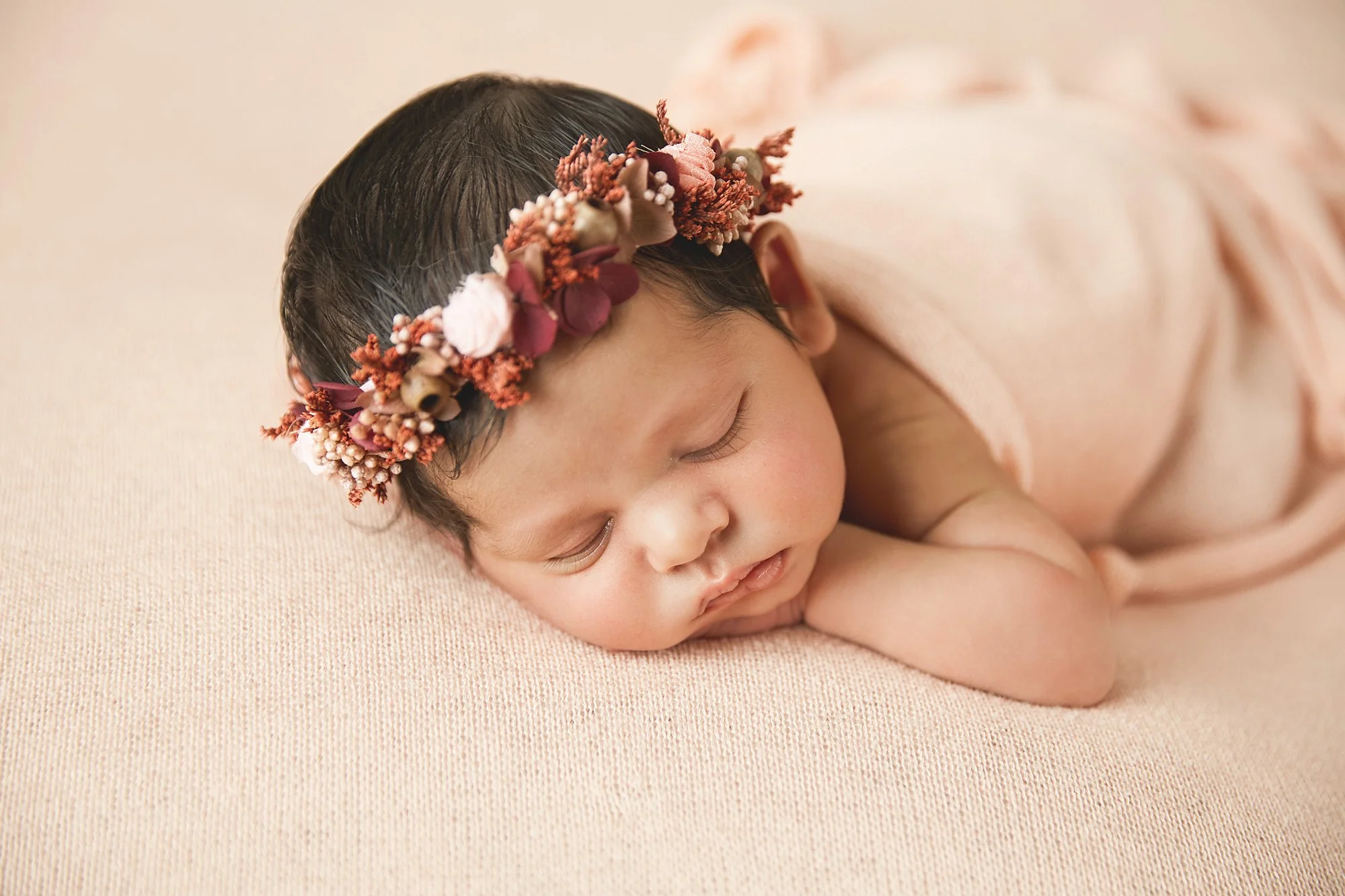 A sleeping baby girl lying on a soft pink surface, wearing a pink dress and a floral headband.