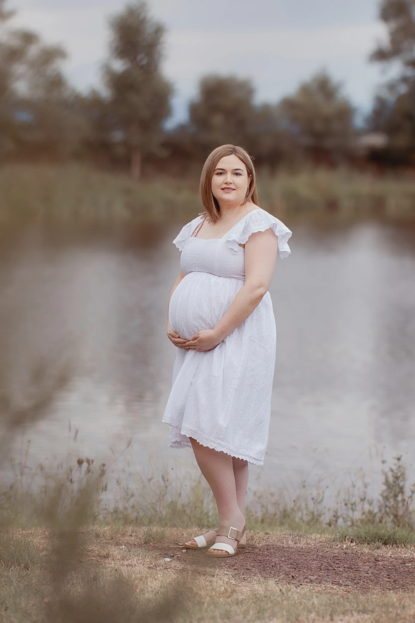 A pregnant woman in a white dress standing outdoors by a lake, looking at the camera, with trees in the background.