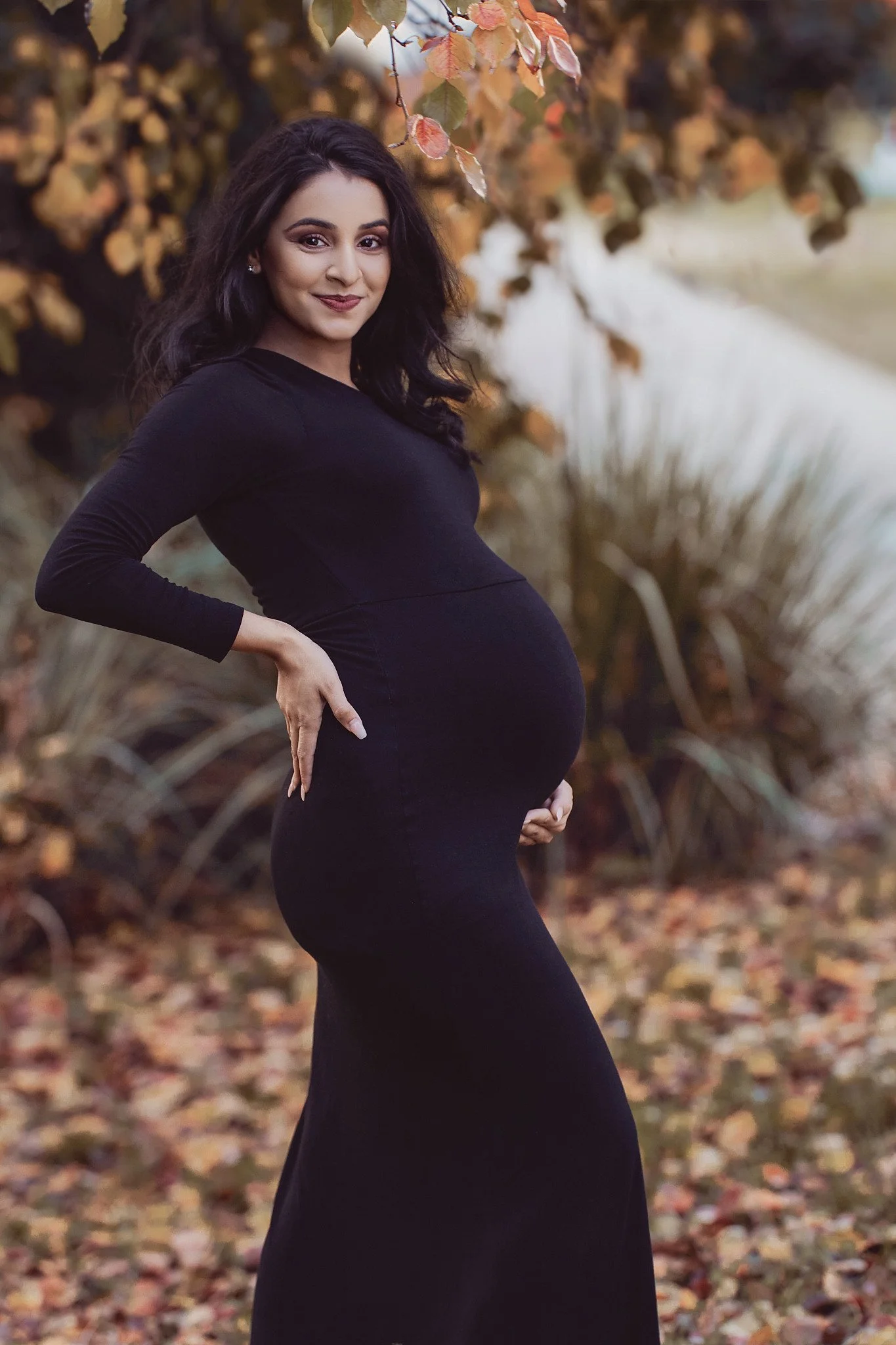 A pregnant woman with long dark hair in a black dress standing outdoors among autumn foliage, smiling and posing with her hand on her hip.
