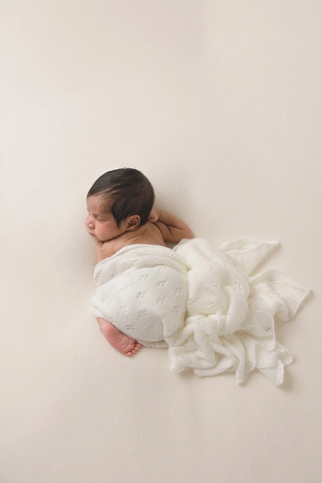 A newborn baby with dark hair sleeping on a cream-colored surface, wrapped in a white knitted blanket.