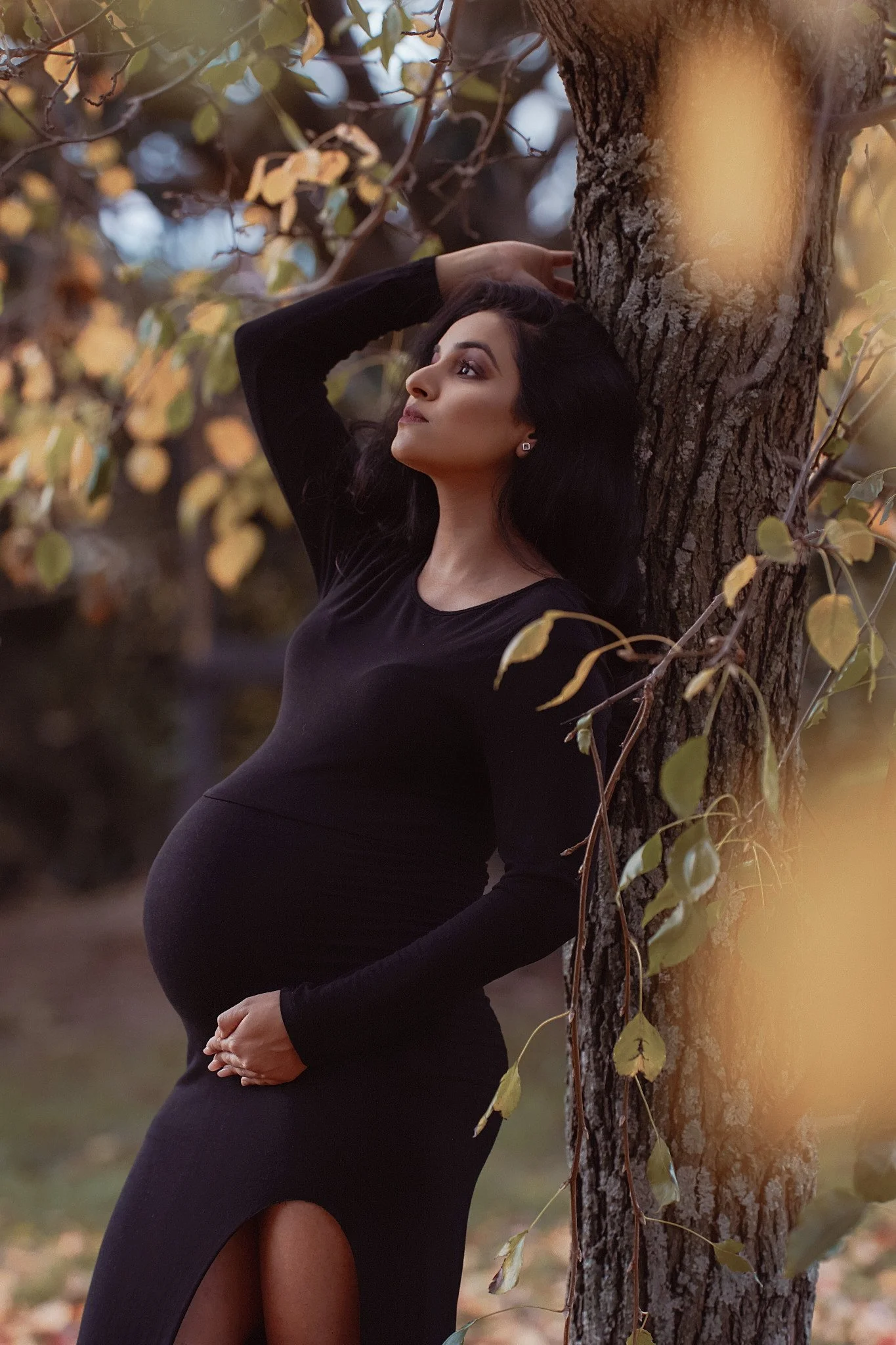 A pregnant woman in a long black dress with a slit, posing outdoors with her back against a tree, looking up and to the side, with her right hand resting on her belly and her left arm raised, touching her hair, surrounded by autumn leaves.