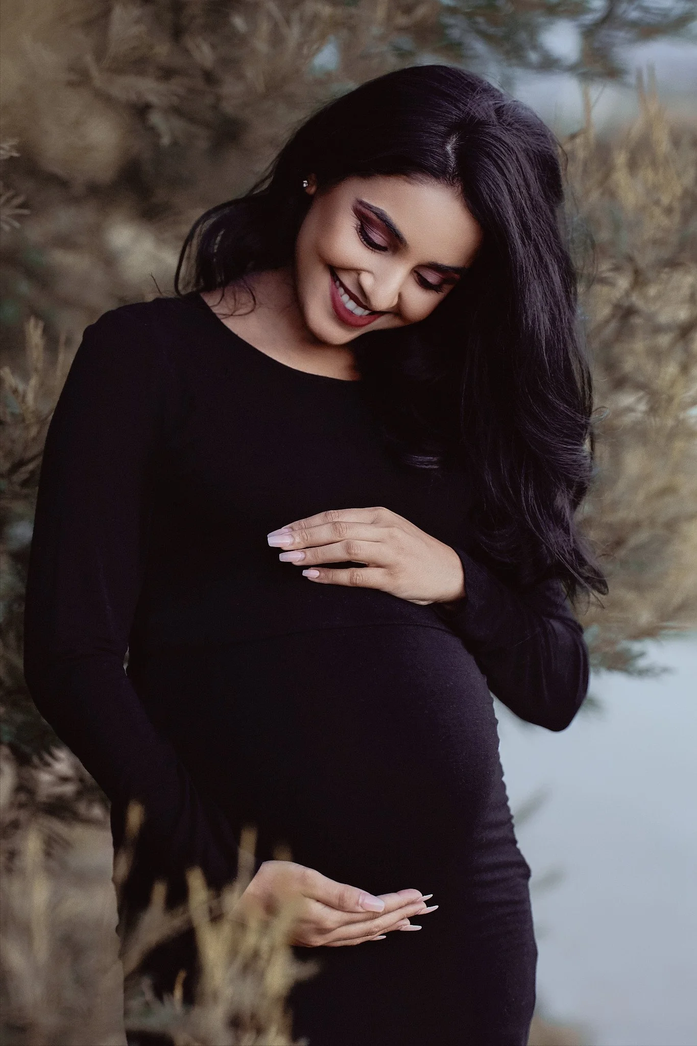 A pregnant woman with long black hair wearing a black dress, smiling and looking down, outdoors with trees and water in the background.