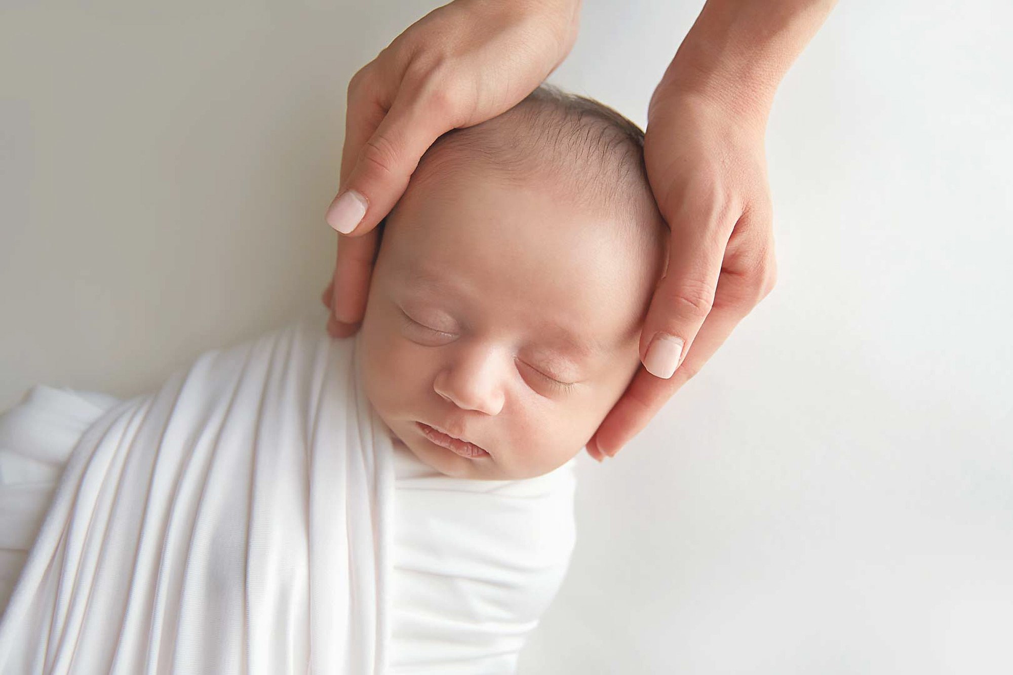 Close-up of a sleeping baby's face with a hand gently holding their head, against a light background.