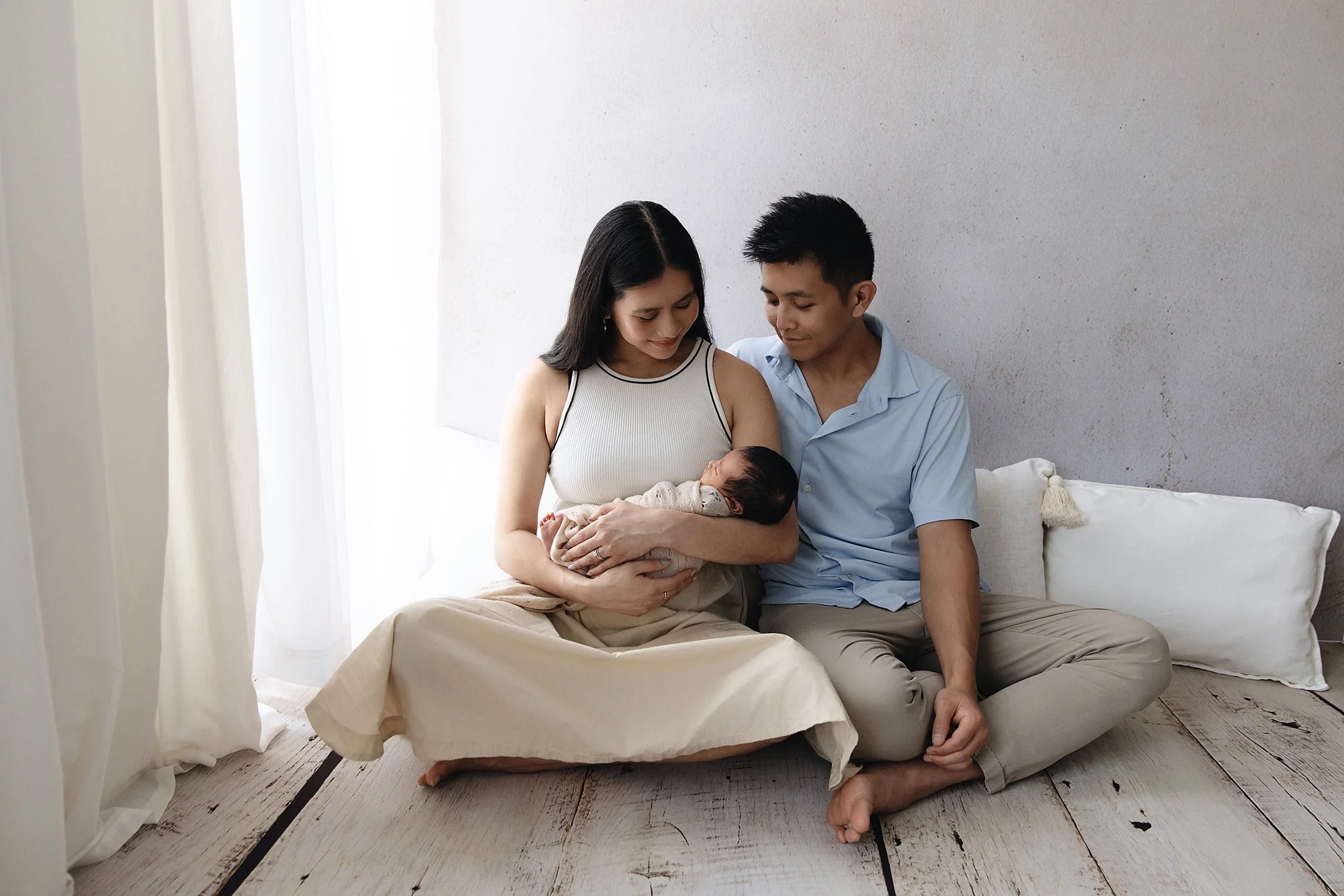 A couple sitting on the floor with their legs crossed, holding a newborn baby. The woman is wearing a sleeveless top and the man a light blue shirt. They are smiling and looking at the baby, with a white wall and curtains in the background.