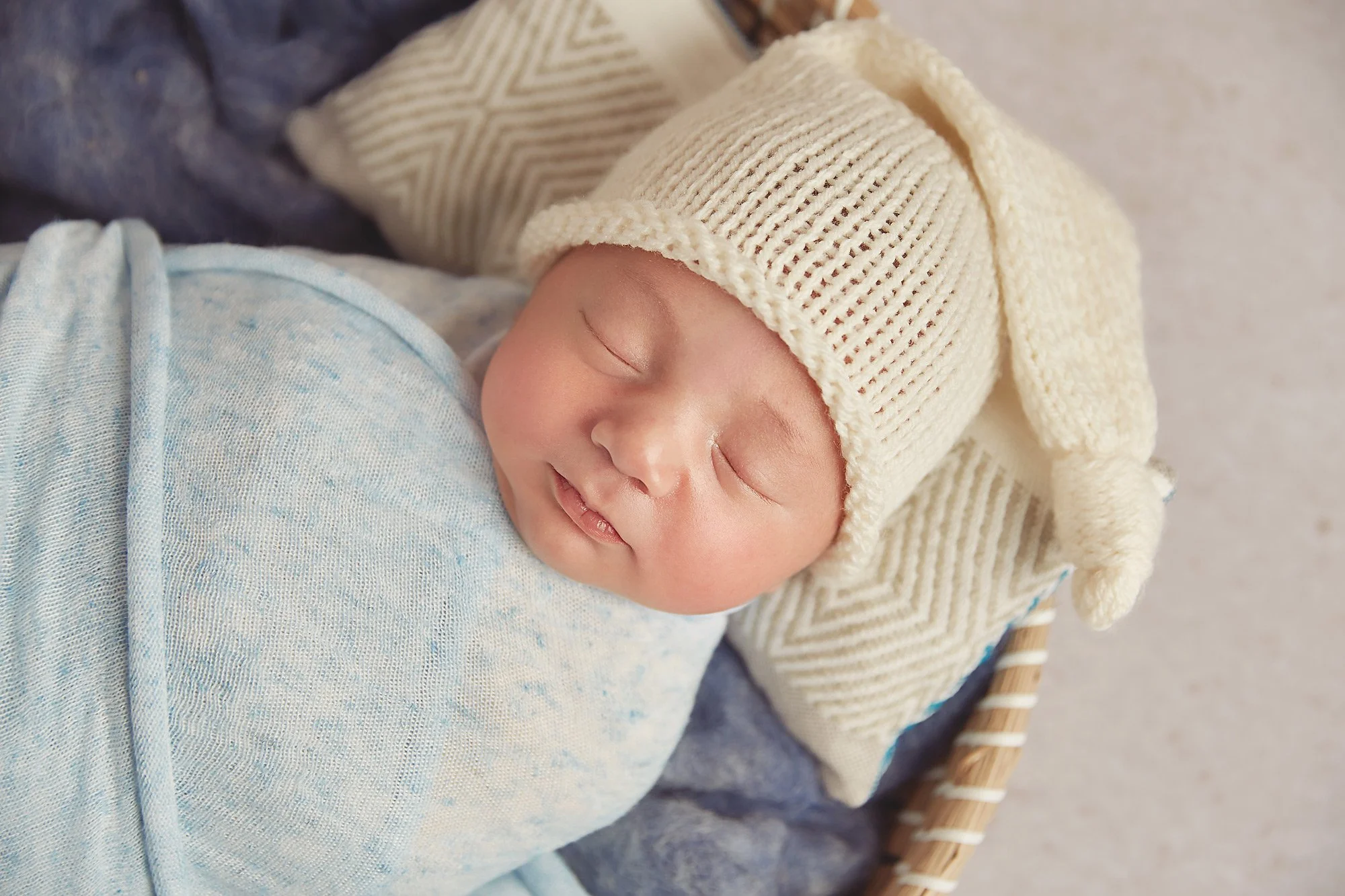 A sleeping baby wrapped in a blue blanket, wearing a beige knit hat with a bunny ear design.