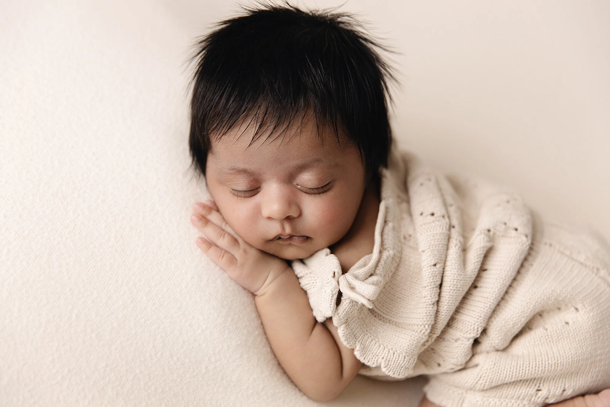 A newborn baby girl with short dark hair and closed eyes, peacefully sleeping with hands tucked under their chin, wearing a cream-colored knitted top with eyelet details, resting on a light beige surface.