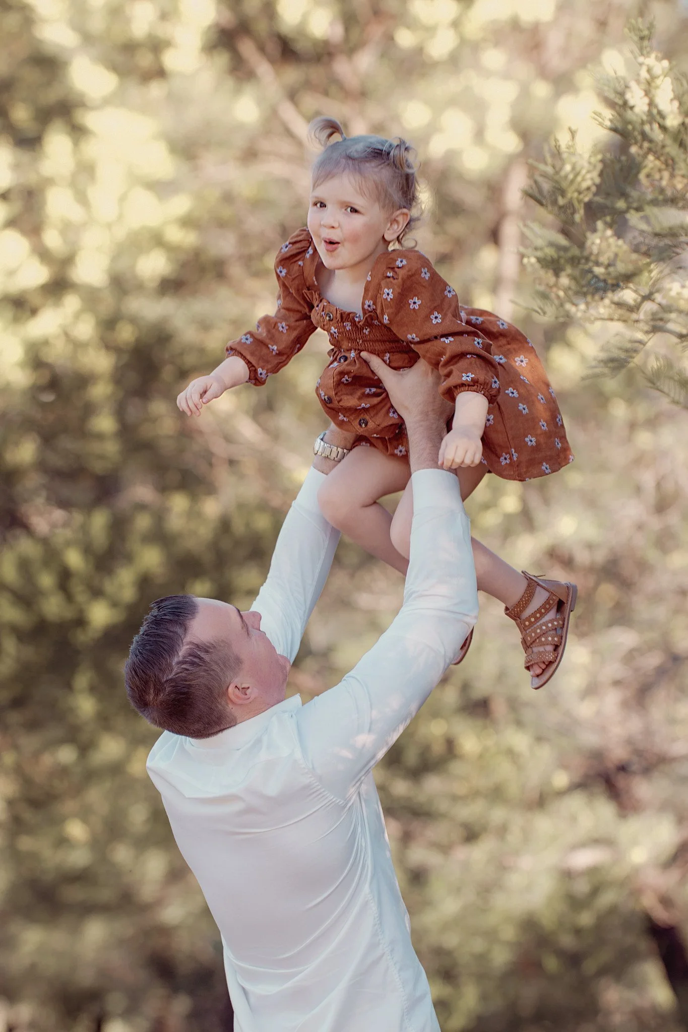 A man lifting a young girl in the air outdoors in a forested area.
