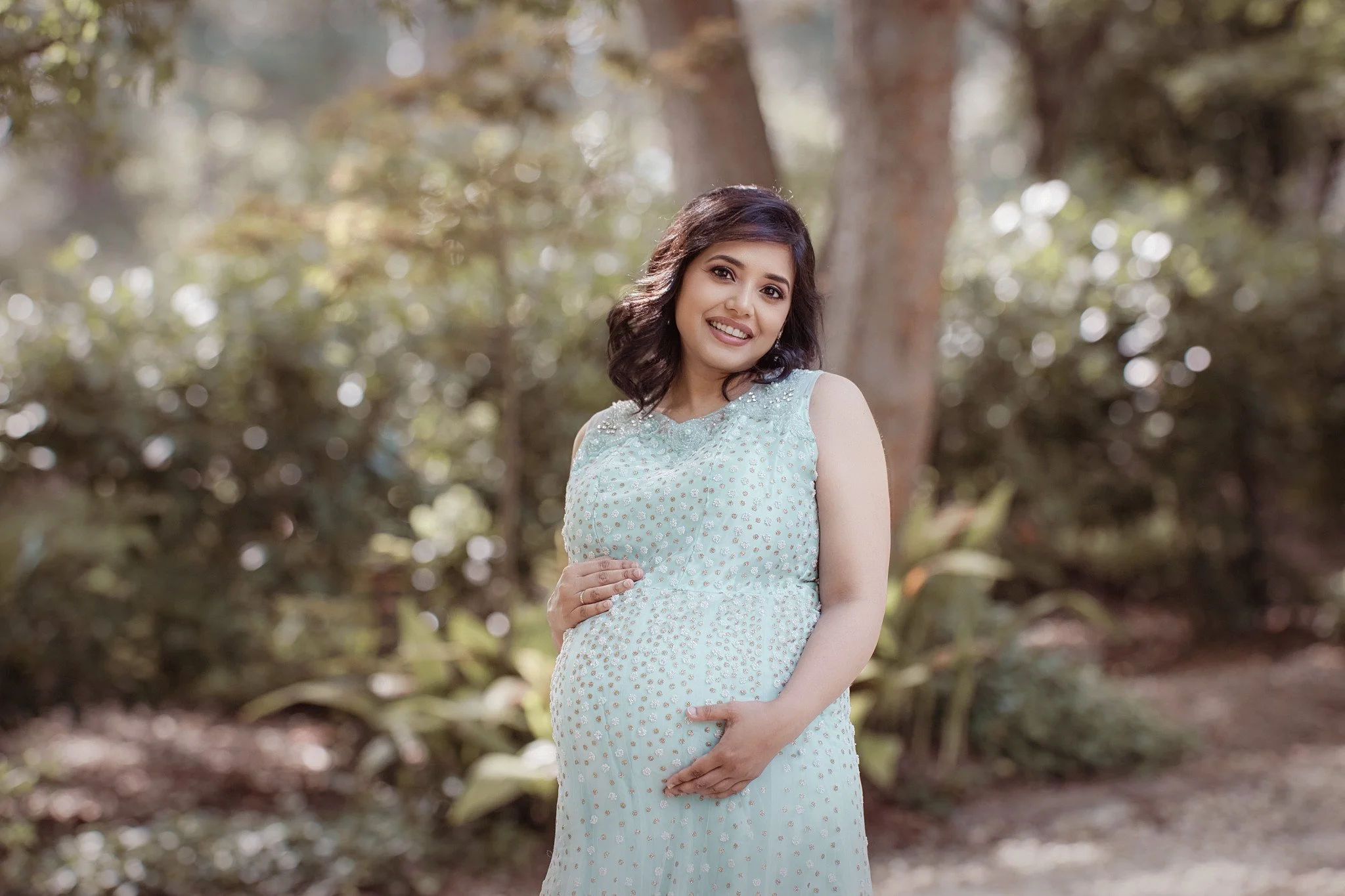 A pregnant woman in a light blue dress standing outdoors in a forested area with trees and greenery, smiling and touching her belly.