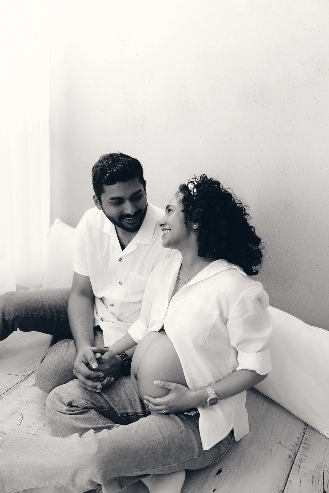 A pregnant woman sits on the floor with her partner, holding her baby bump and smiling at each other in a cozy room.