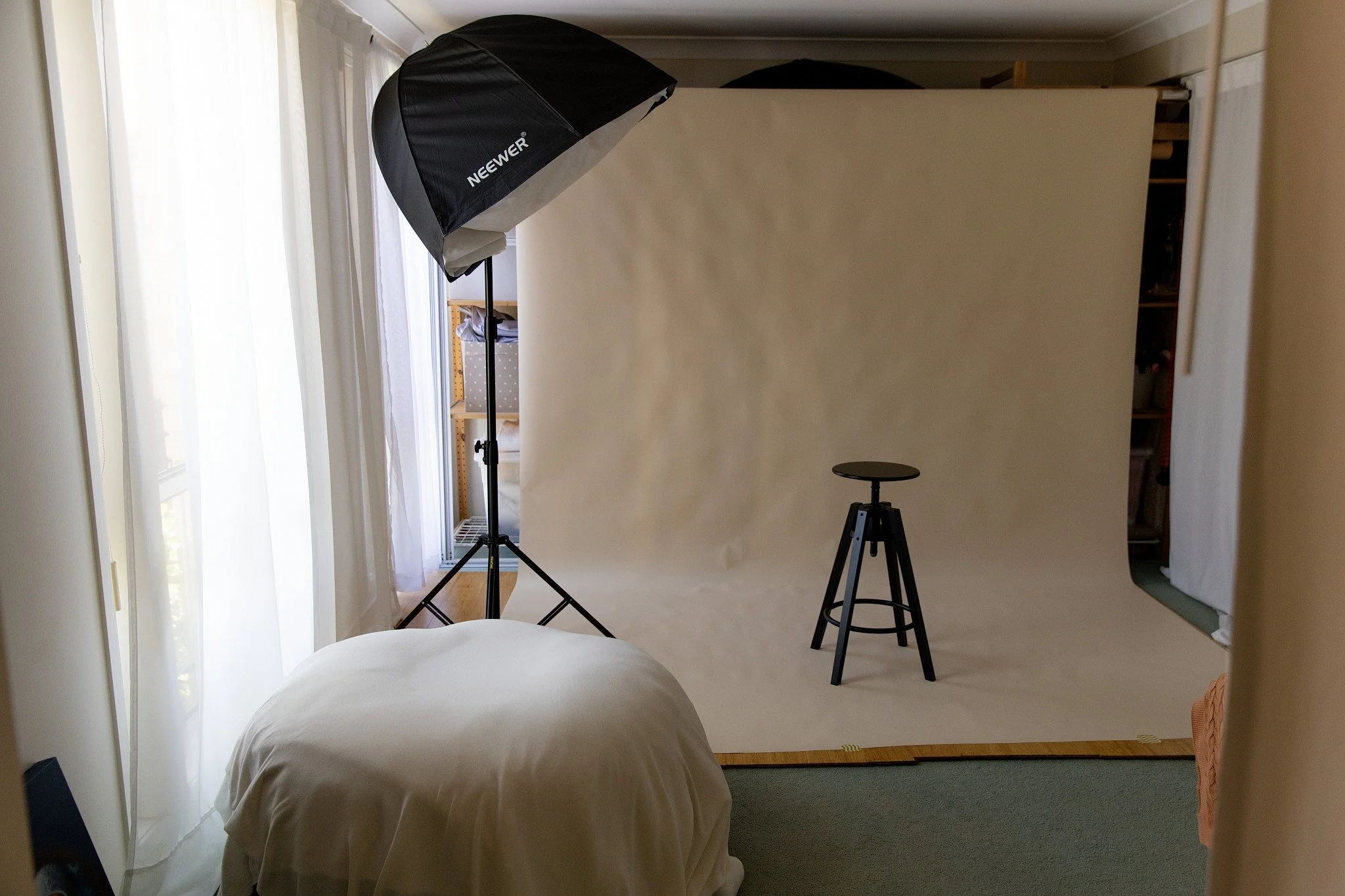 A photography studio setup with a cream backdrop, a photography light box, and a tripod, located in a room with white curtains and a beanbag.