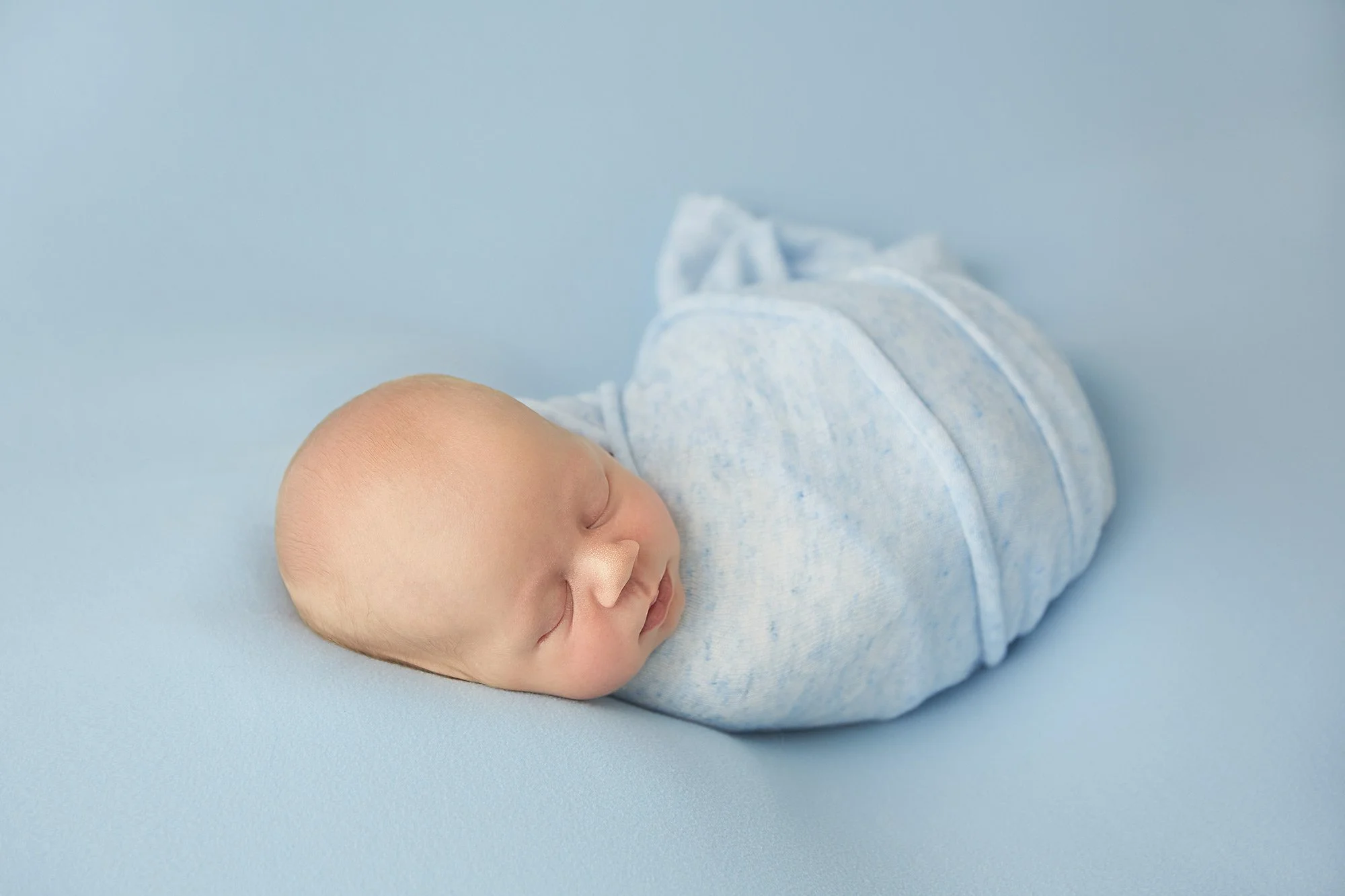 A sleeping newborn baby wrapped in a light blue blanket on a matching light blue surface.